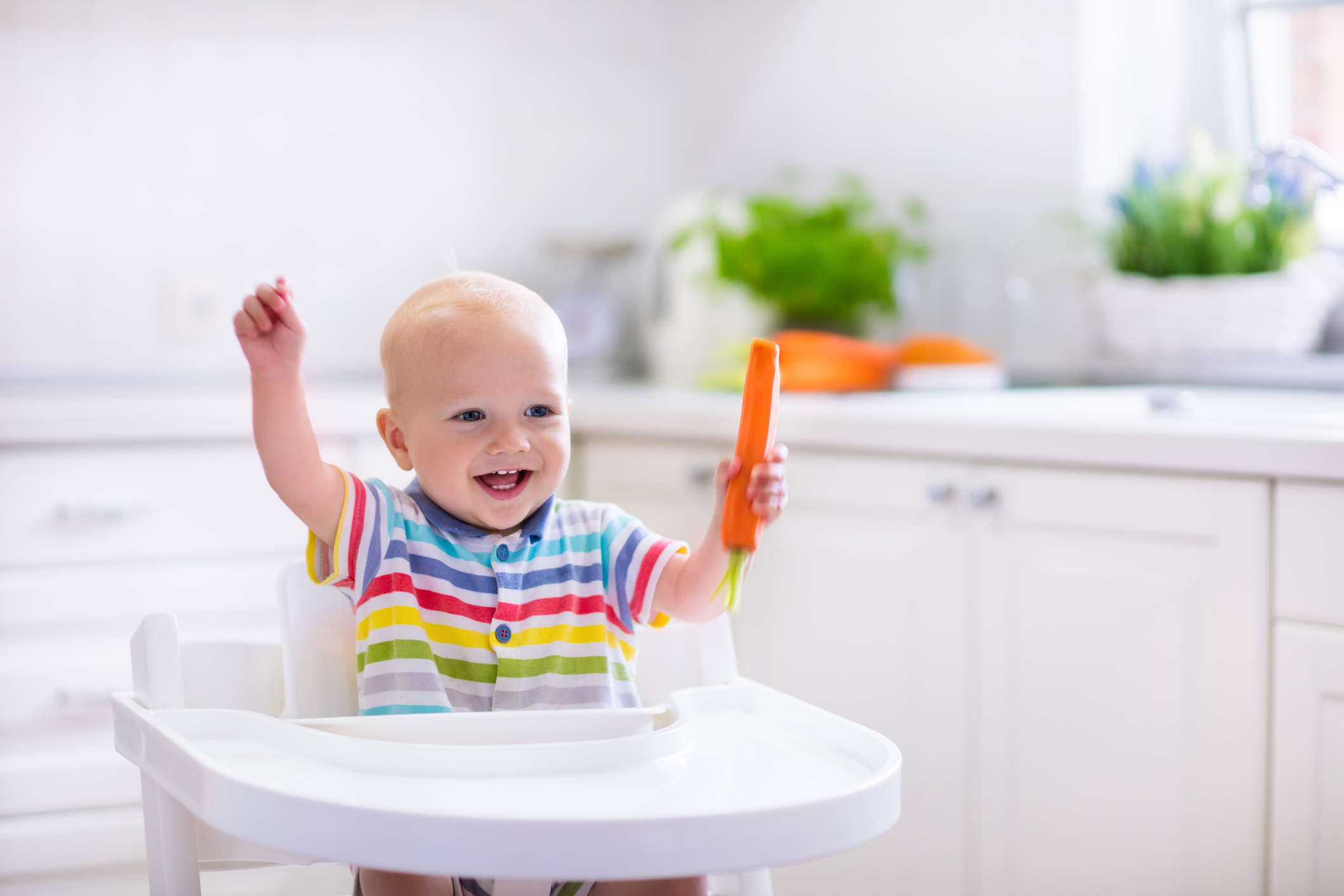 Happy baby sitting in high chair eating carrot in a white kitchen (Thinkstock/PA)