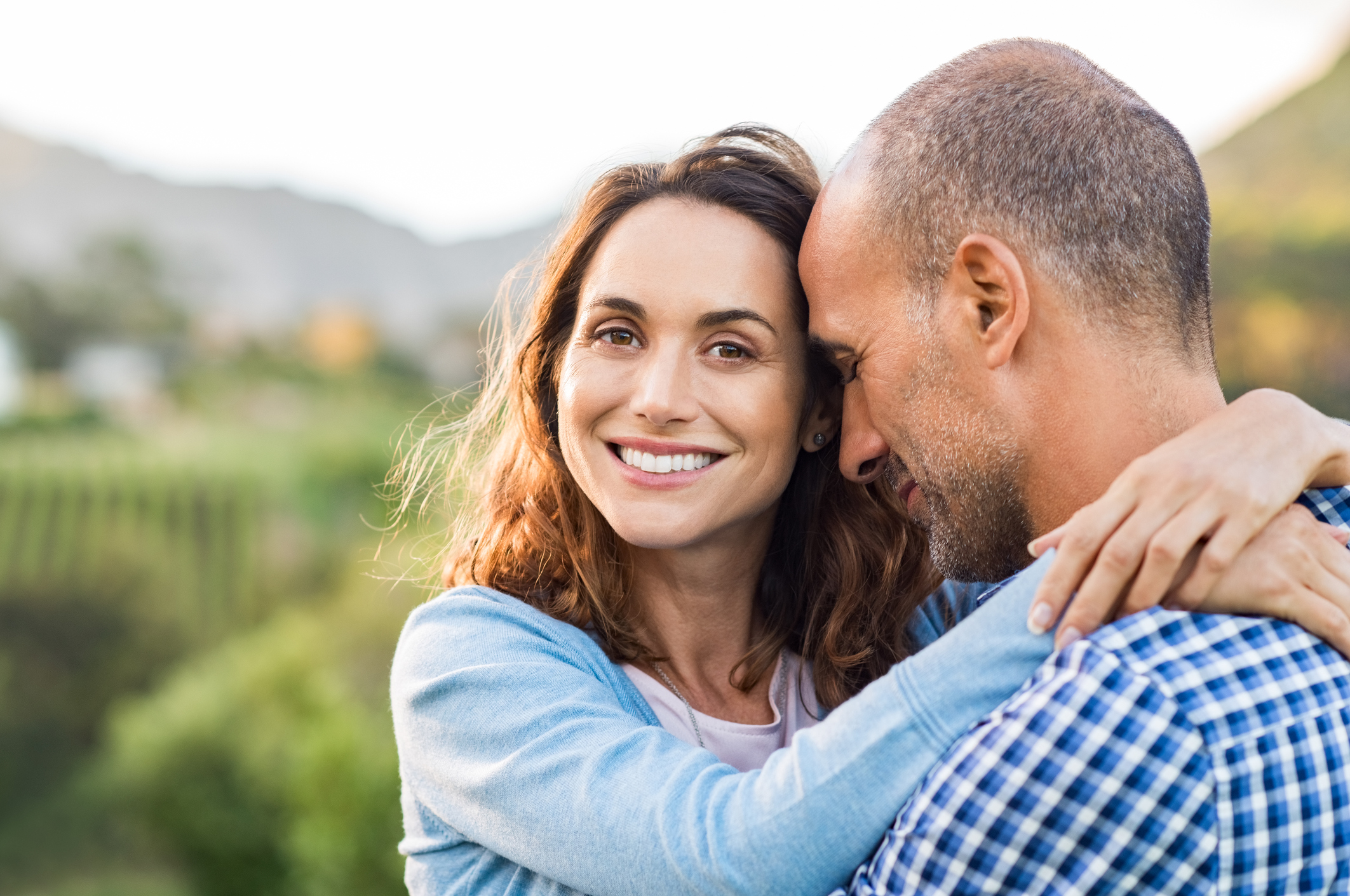 Mature romantic couple embracing outdoor. Happy woman embracing her multiethnic boyfriend at park during sunset. Smiling brunette woman in love with her husband looking at camera.
