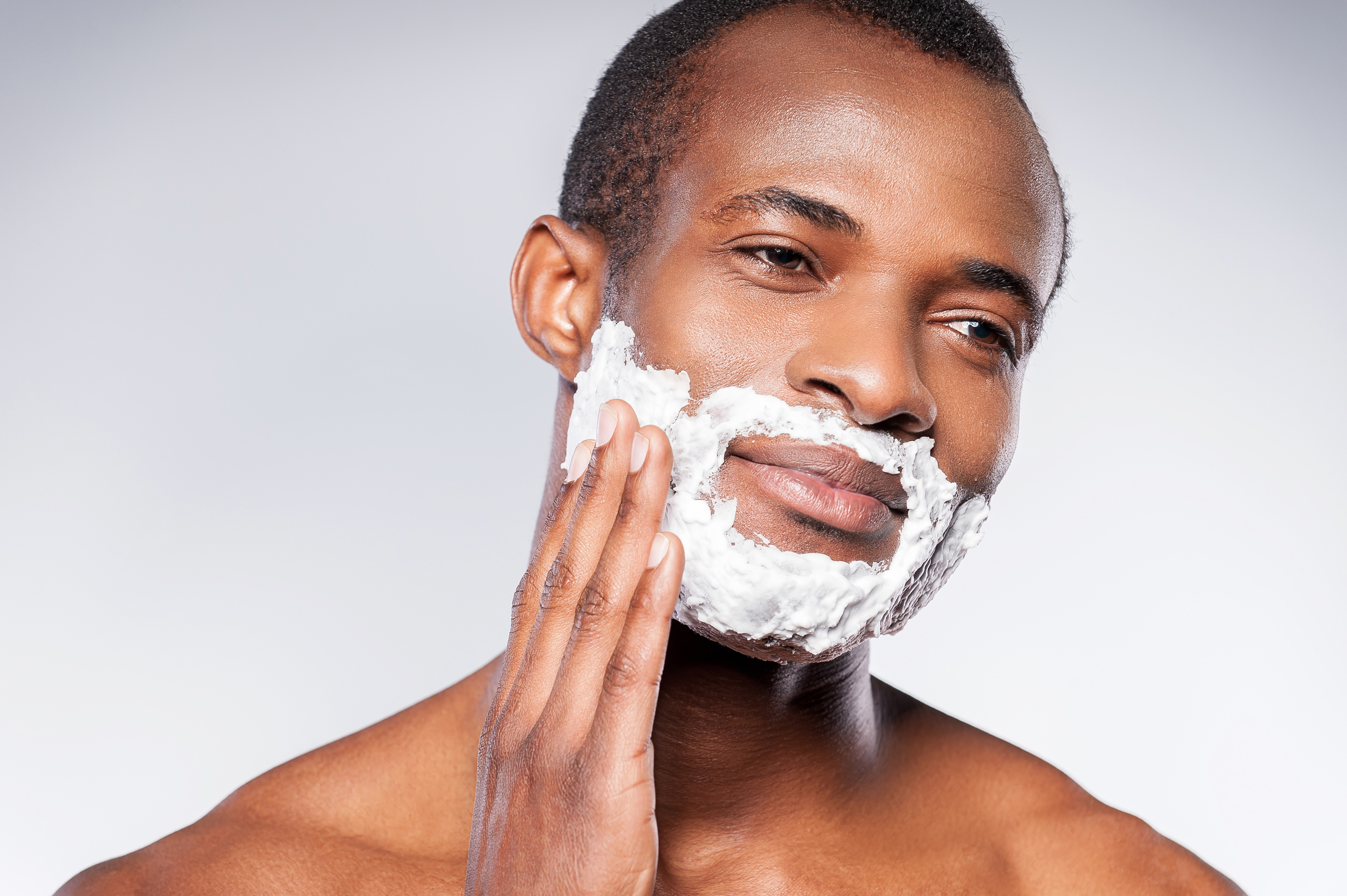 Handsome shirtless African man shaving his face and looking at camera while standing against grey background