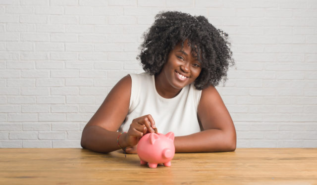 Young african american woman sitting on the table holding piggy bank with a happy face standing and smiling with a confident smile showing teeth