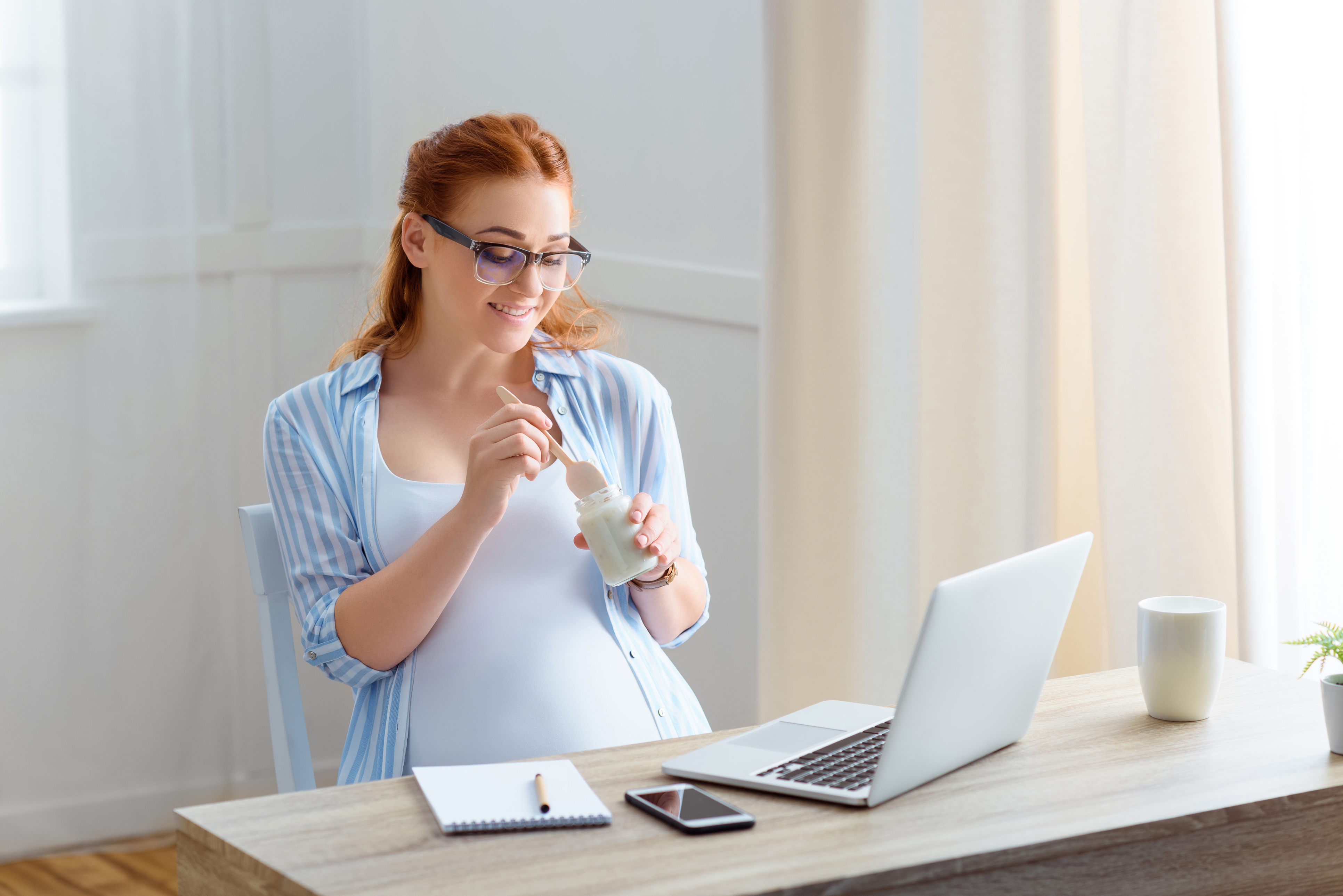 smiling redhead pregnant woman eating yoghurt while sitting at table with laptop, notepad and smartphone at home
