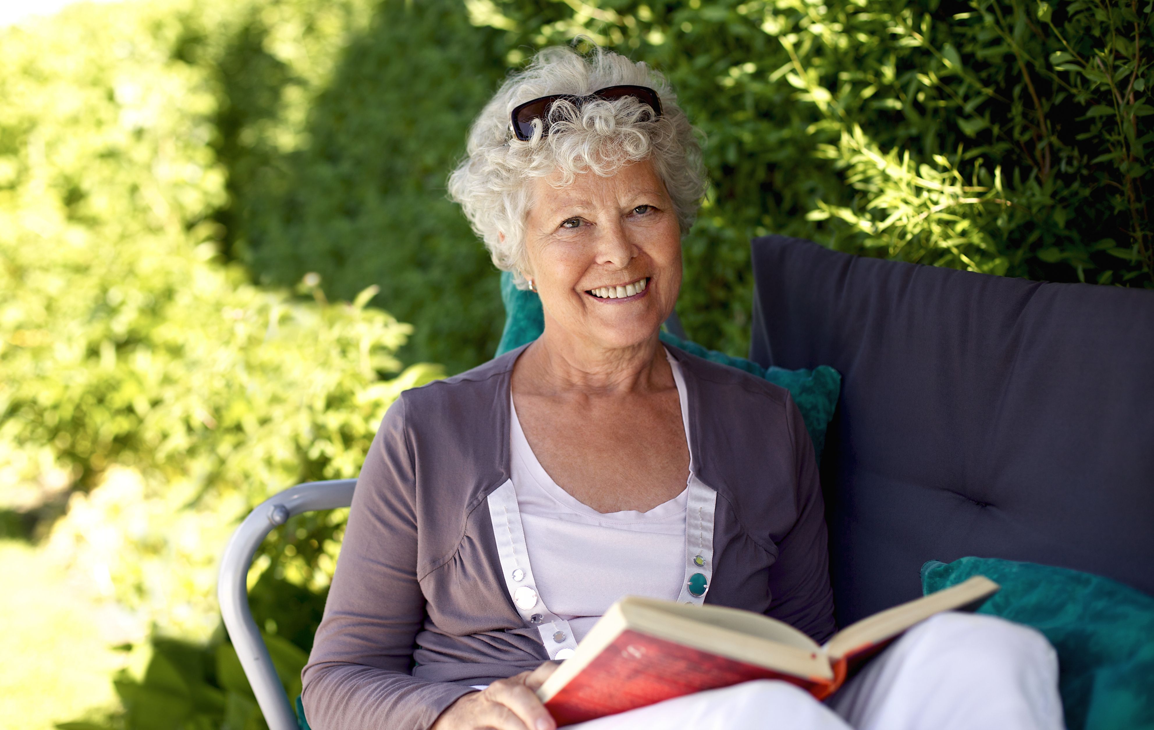 Elder woman sitting on a chair in backyard garden holding a book and looking at camera smiling