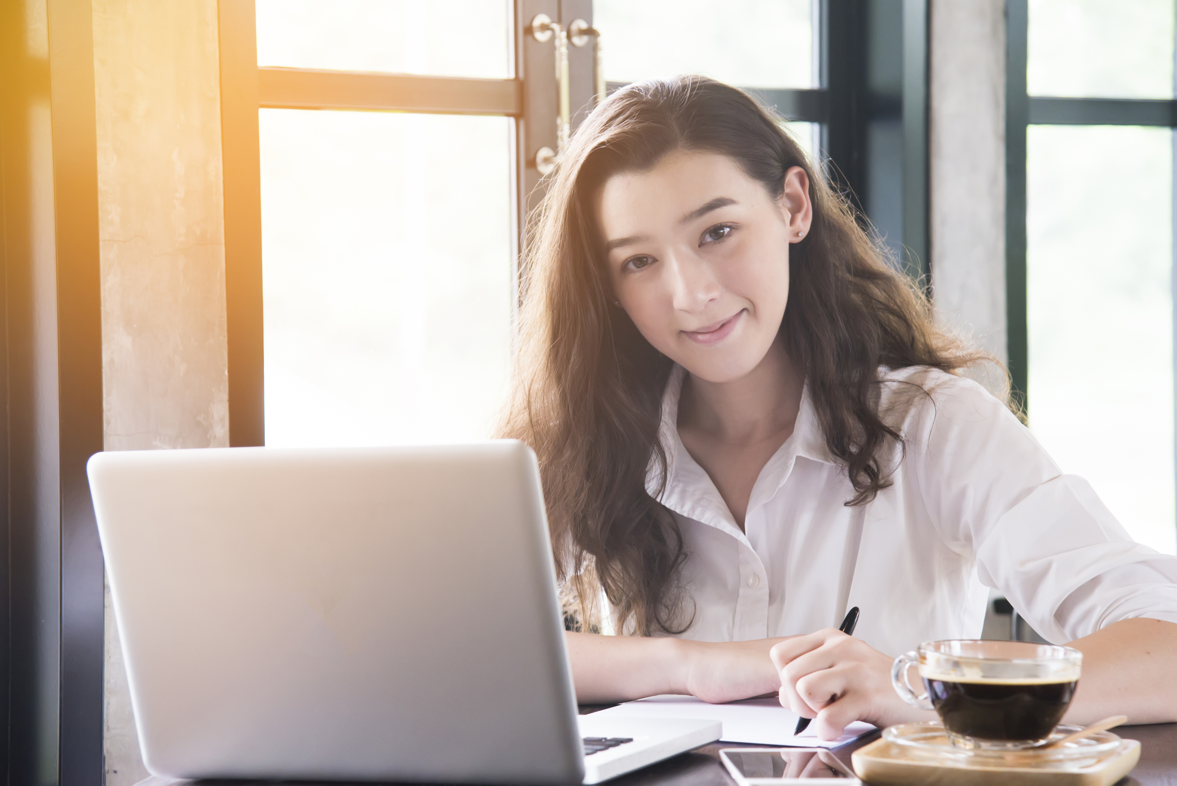 Young woman sitting in coffee shop at wooden table, drinking coffee and using smartphone.On table is laptop for internet shopping or working online.
