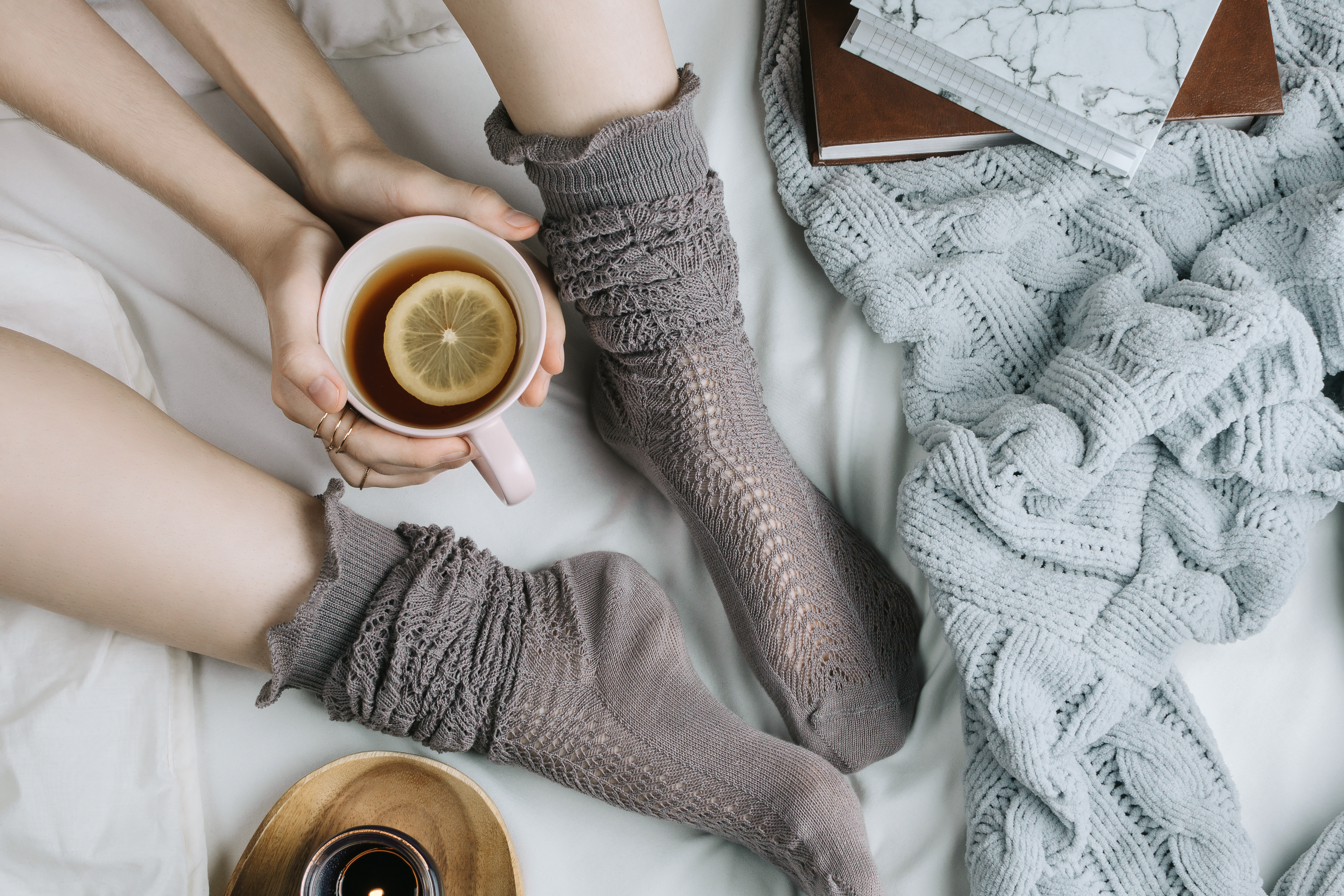 Cozy flatlay of woman's legs in warm grey stockings in bed with knitted sweater and books holding cup of lemon tea, selective focus