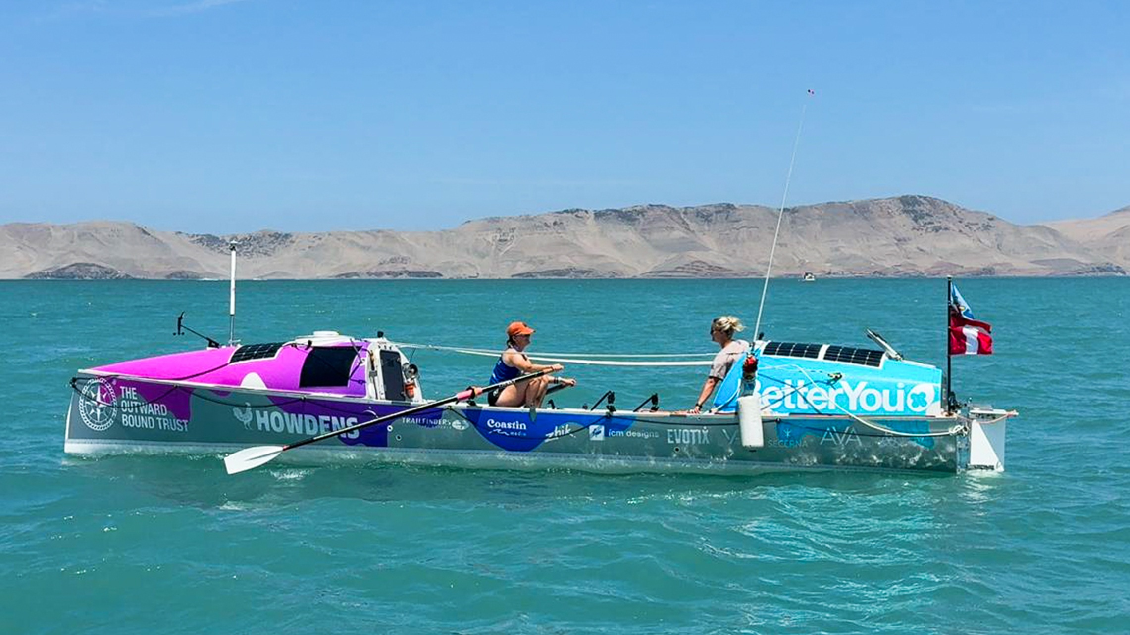 Two women in a multicoloured boat on the sea with a pale blue sky in the background