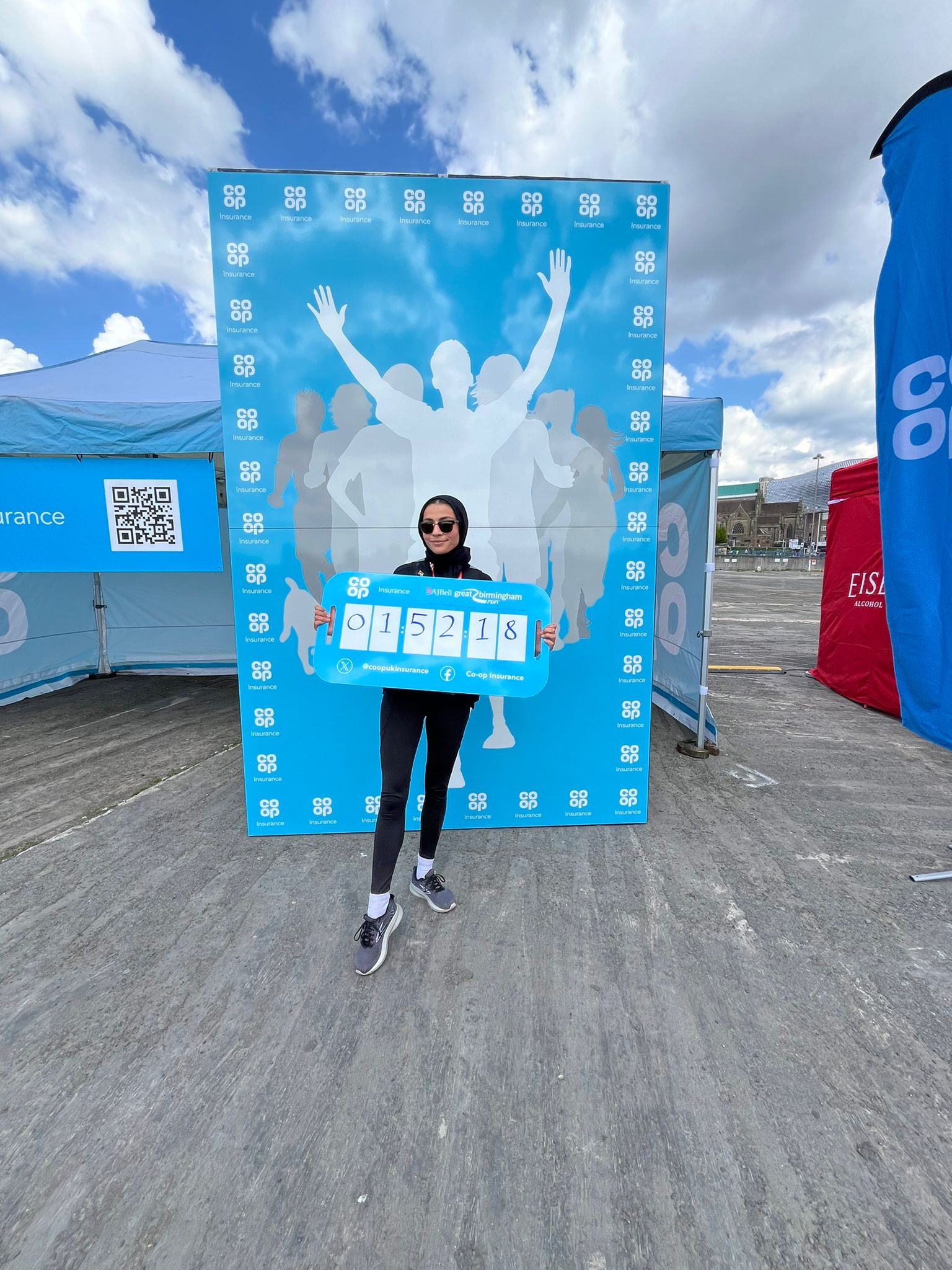 Tanzeela Khalid holding a bib number in front of a running event sign with Coop branding