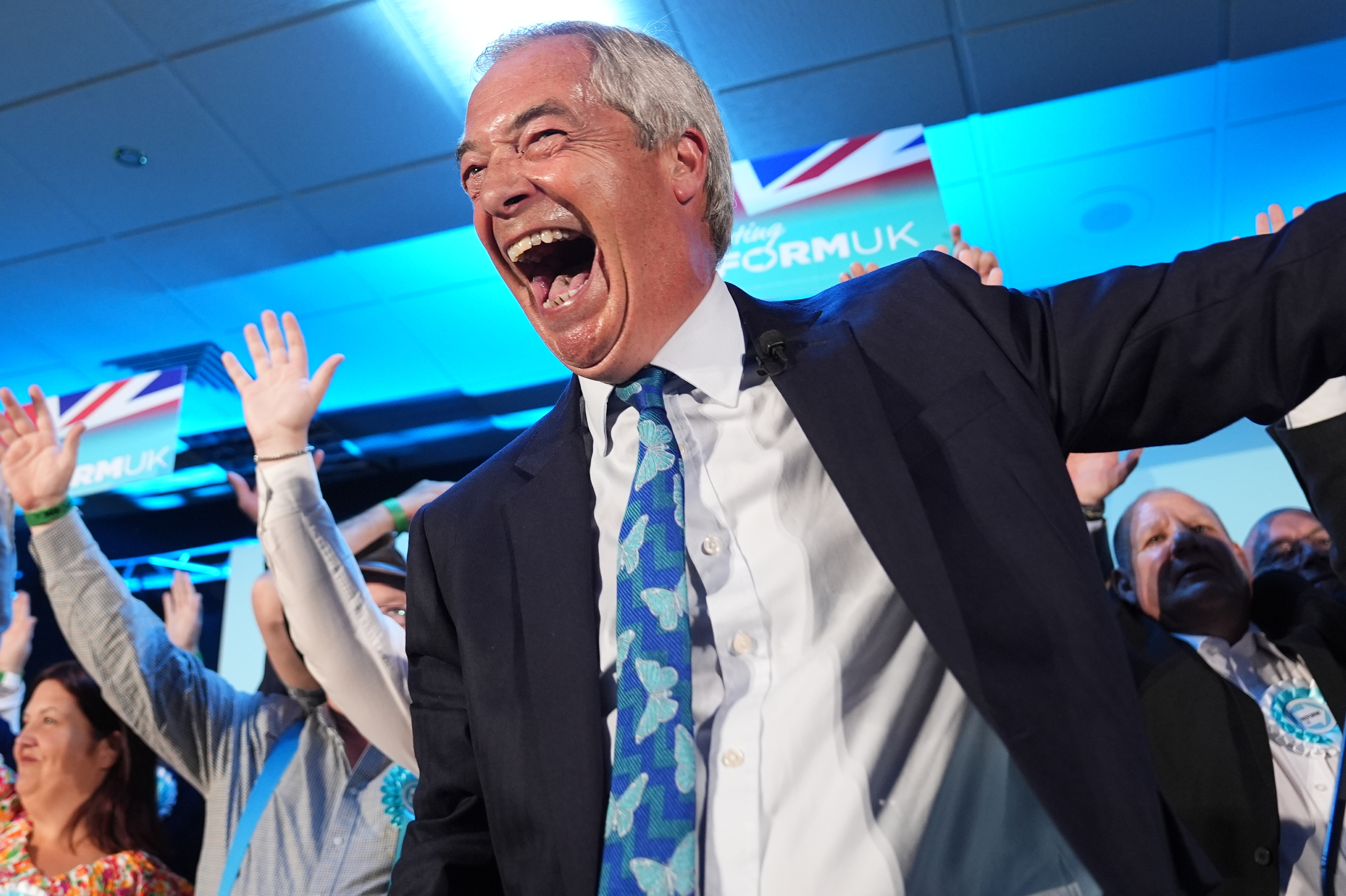 Nigel Farage smiling while celebrating, in front of Reform supporters with their arms in the air