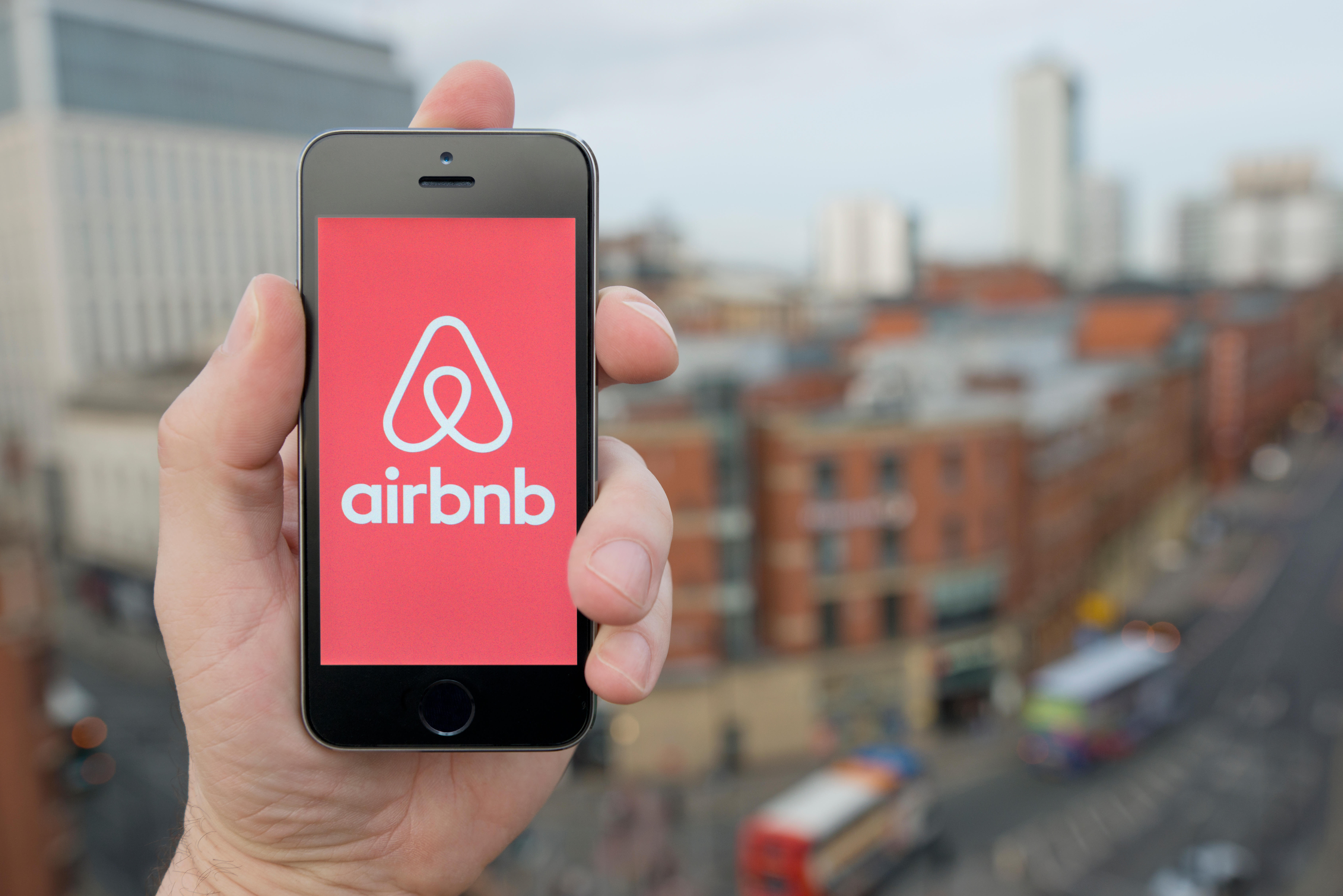 A hand holding a mobile phone with the Airbnb logo on it up above a overlooking a busy city street