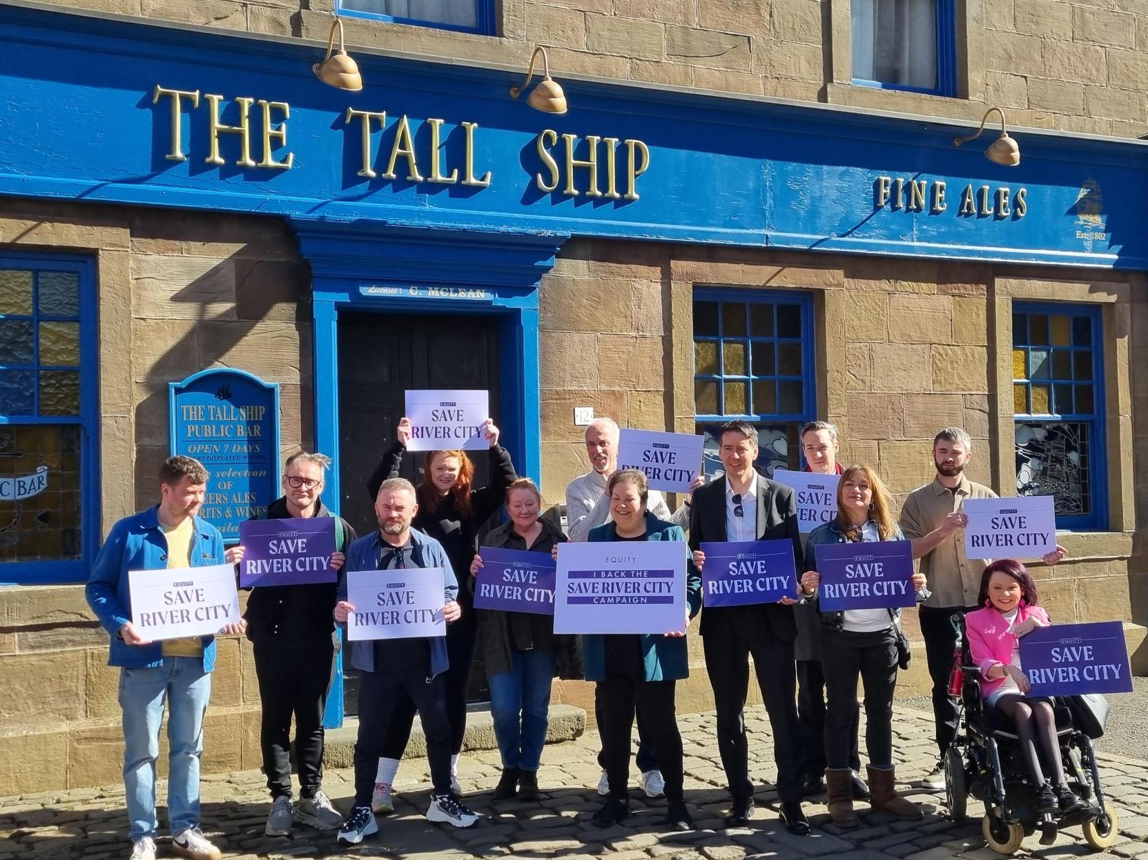 A group of River City stars and politicians holding 'Save River City' signs outside the soap's The Tall Ship pub