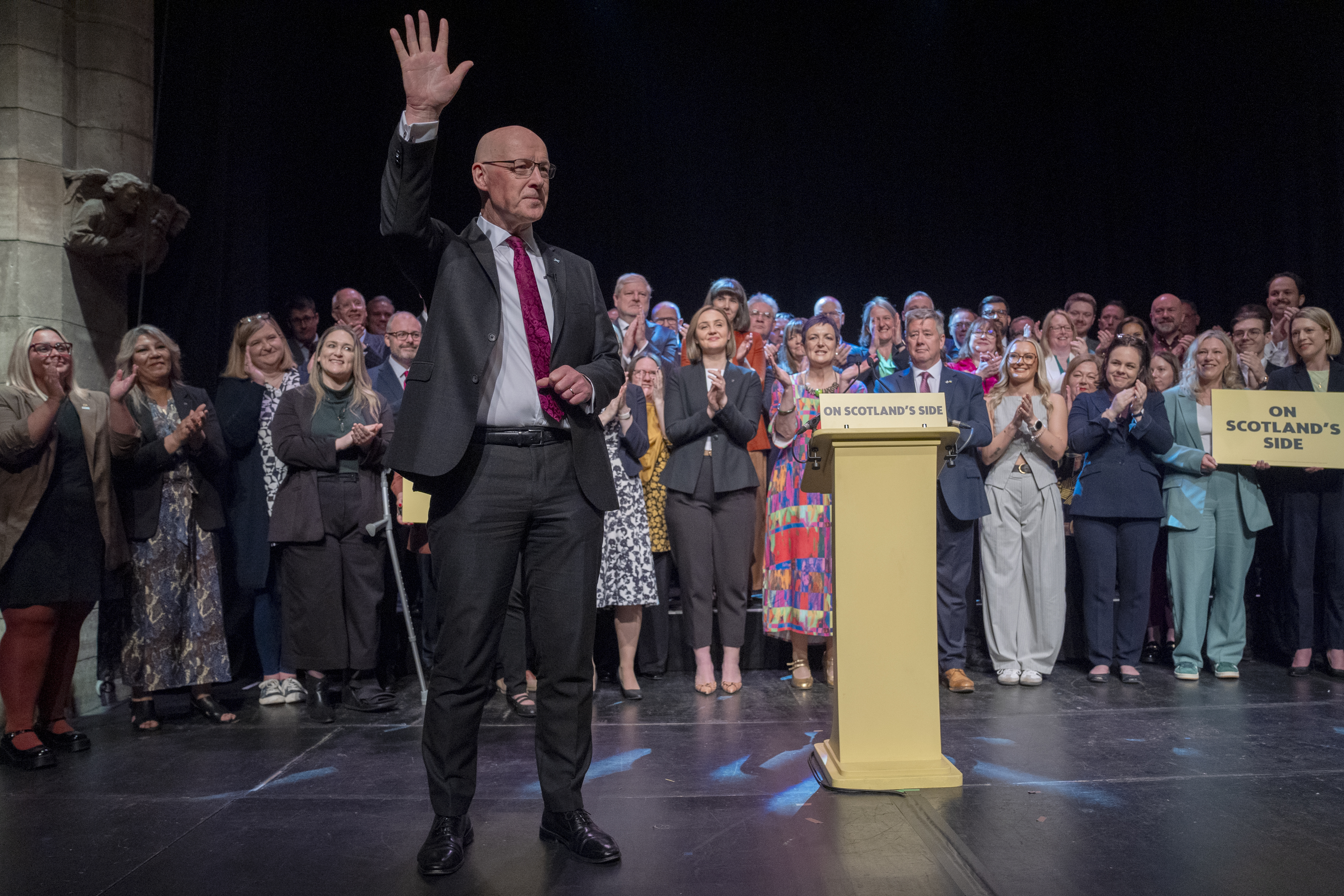 John Swinney waving on a stage, with SNP members and politicians behind him
