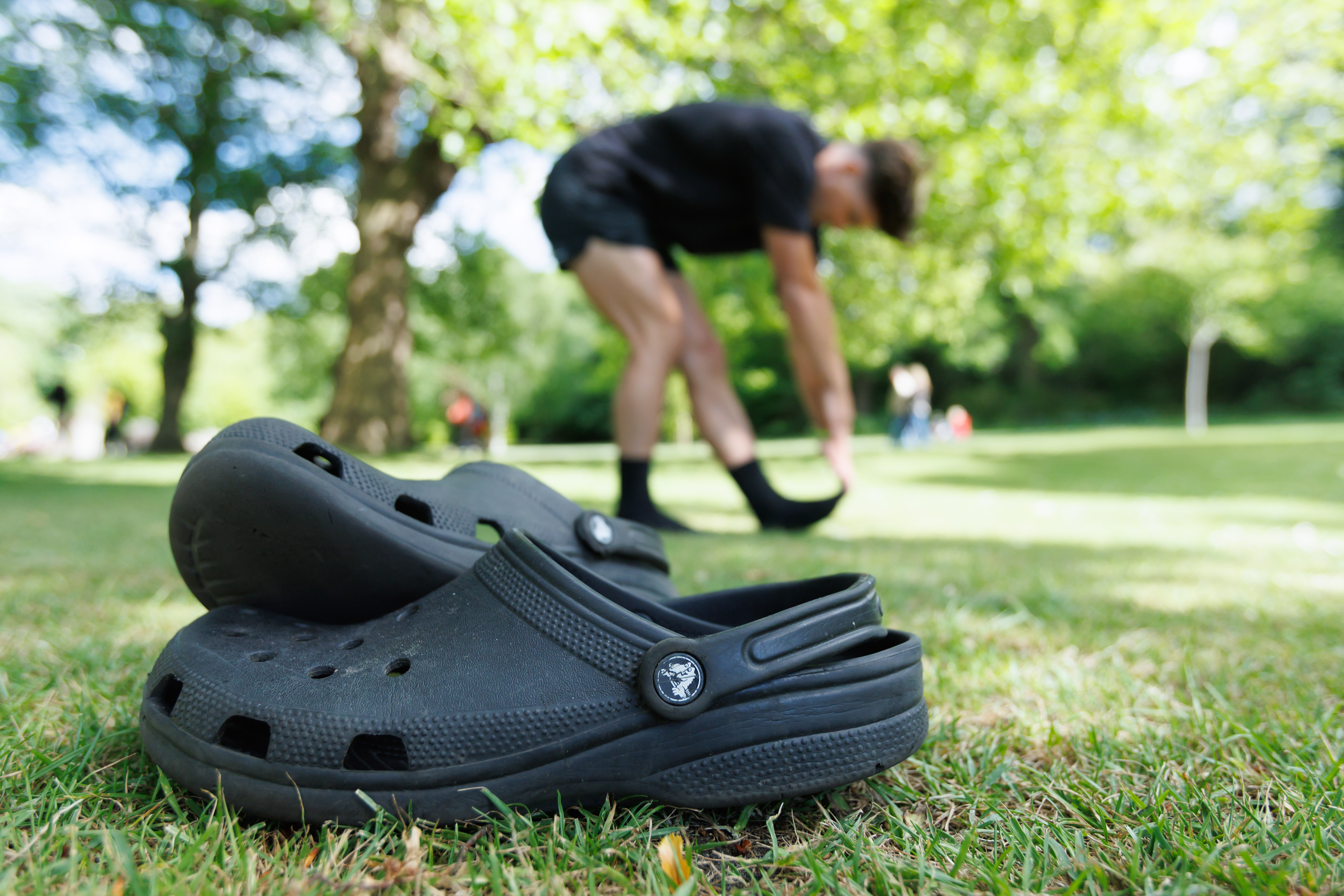 A close-up view of a pair of black Crocs on grass, with Danny Campbell in the background doing some stretches