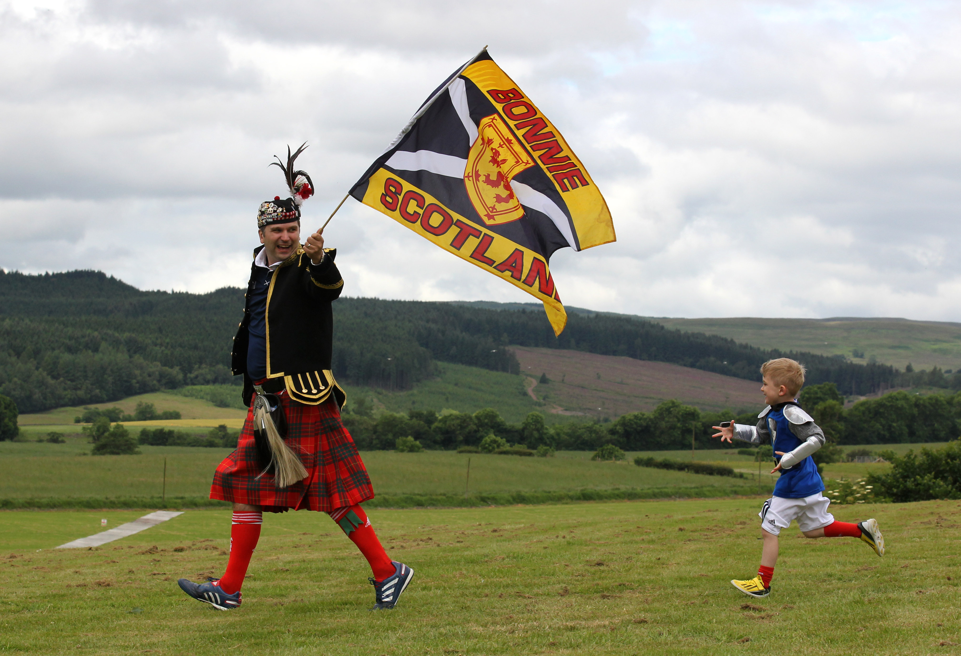 A young boy chases a man wearing a kilt who is holding a Scottish flag