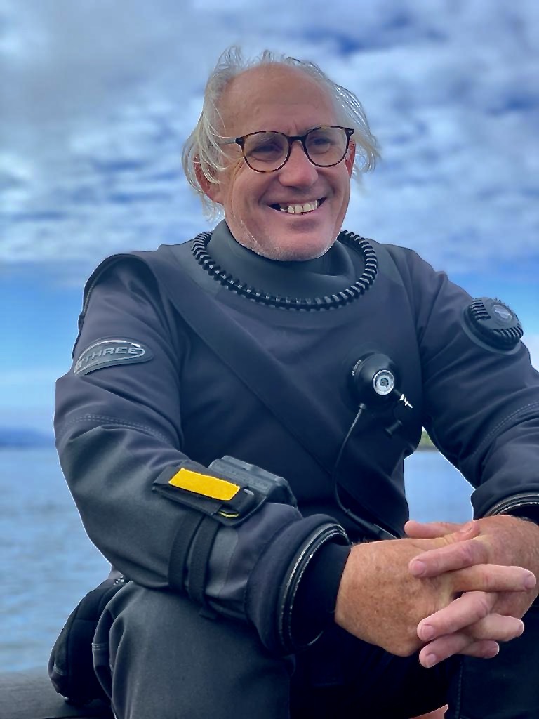 Graeme Bruce smiling while wearing diving gear, with the sea behind him
