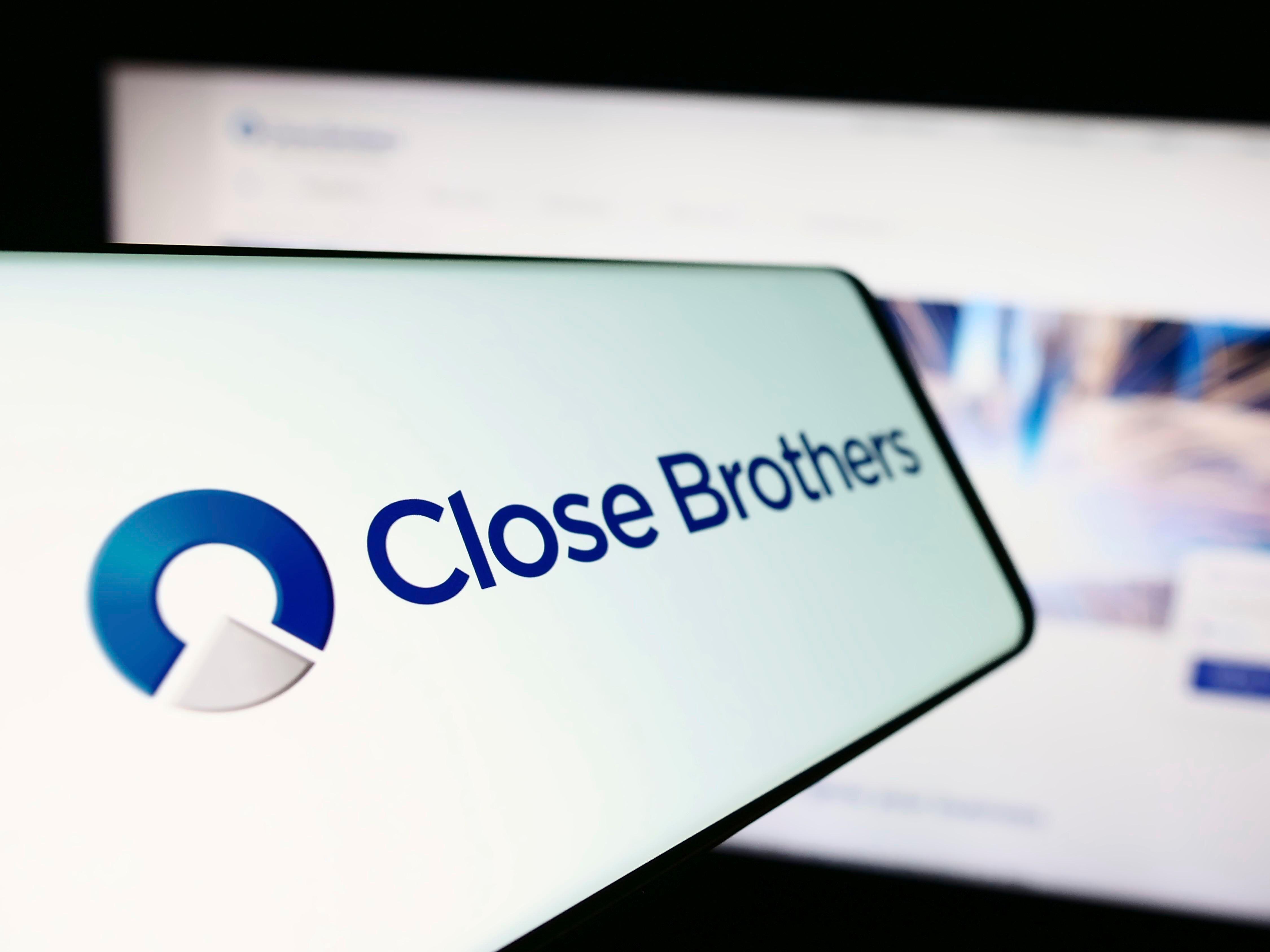 Mobile phone with logo of Close Brothers Group in front of its website displayed in the background on a monitor