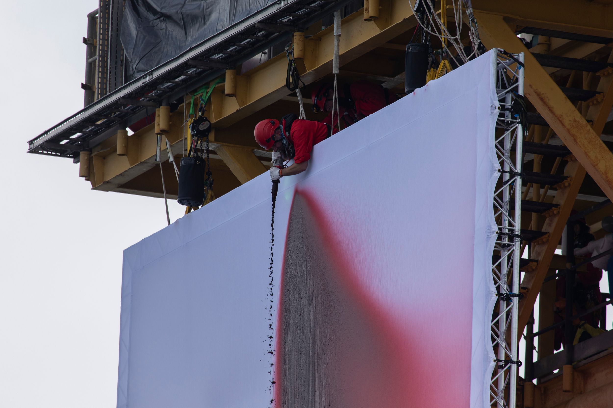 A man in red T-shirt and hardhat sprays red liquid on a white canvas