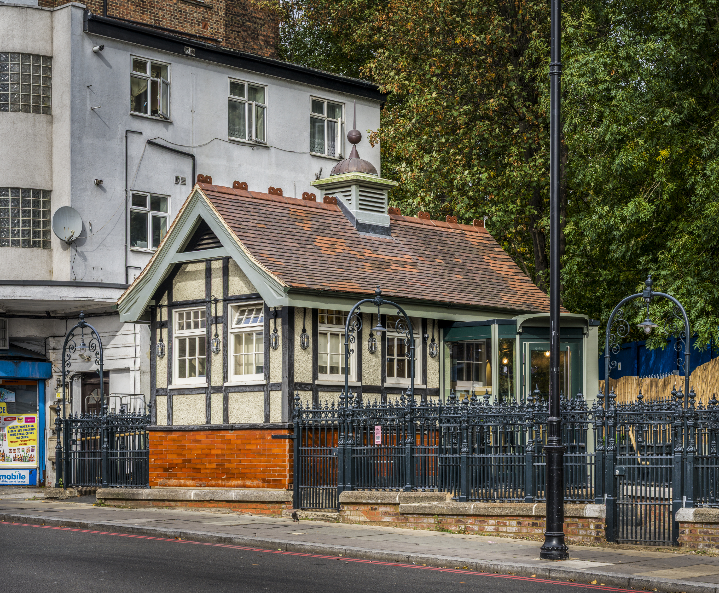 A mock-Tudor small Victorian building surrounded by metal railings on the side of a road, with other buildings and trees behind