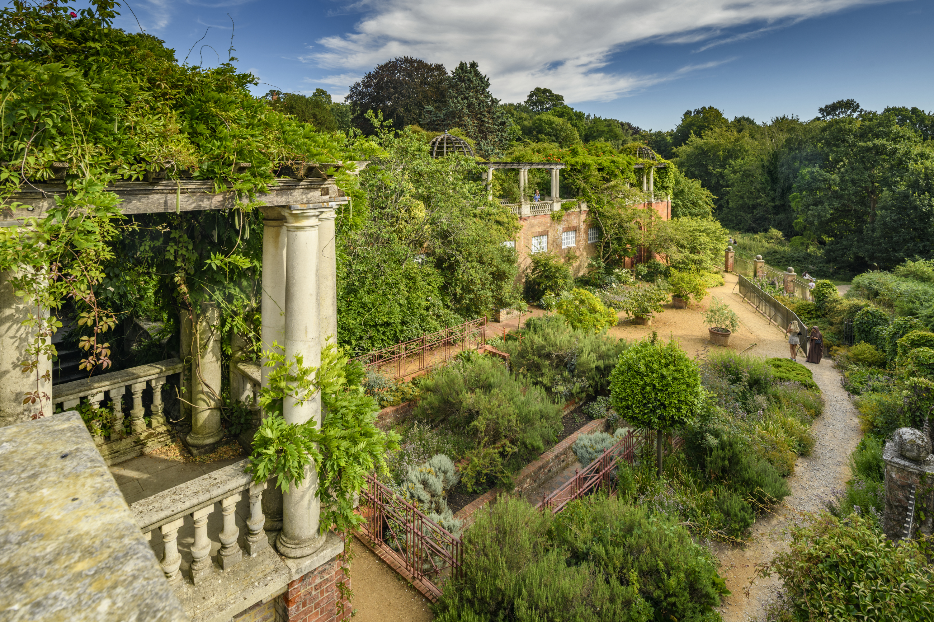 Image shows a garden with largescale stone, brick and metal pergola overgrown with vegetation