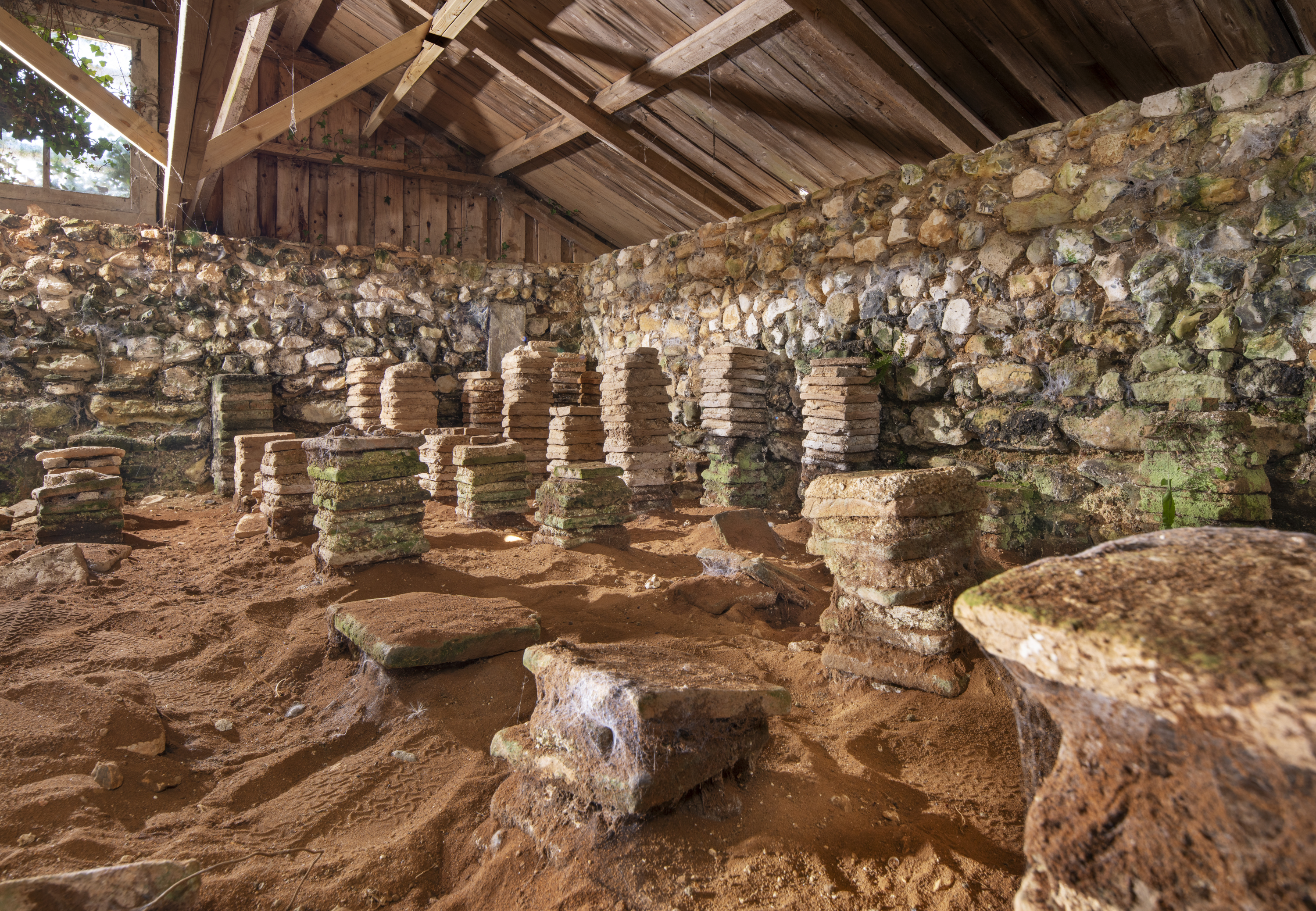 Brick pillars creating a Roman hypocaust on a sandy floor are sheltered by a stone and timber roofed building