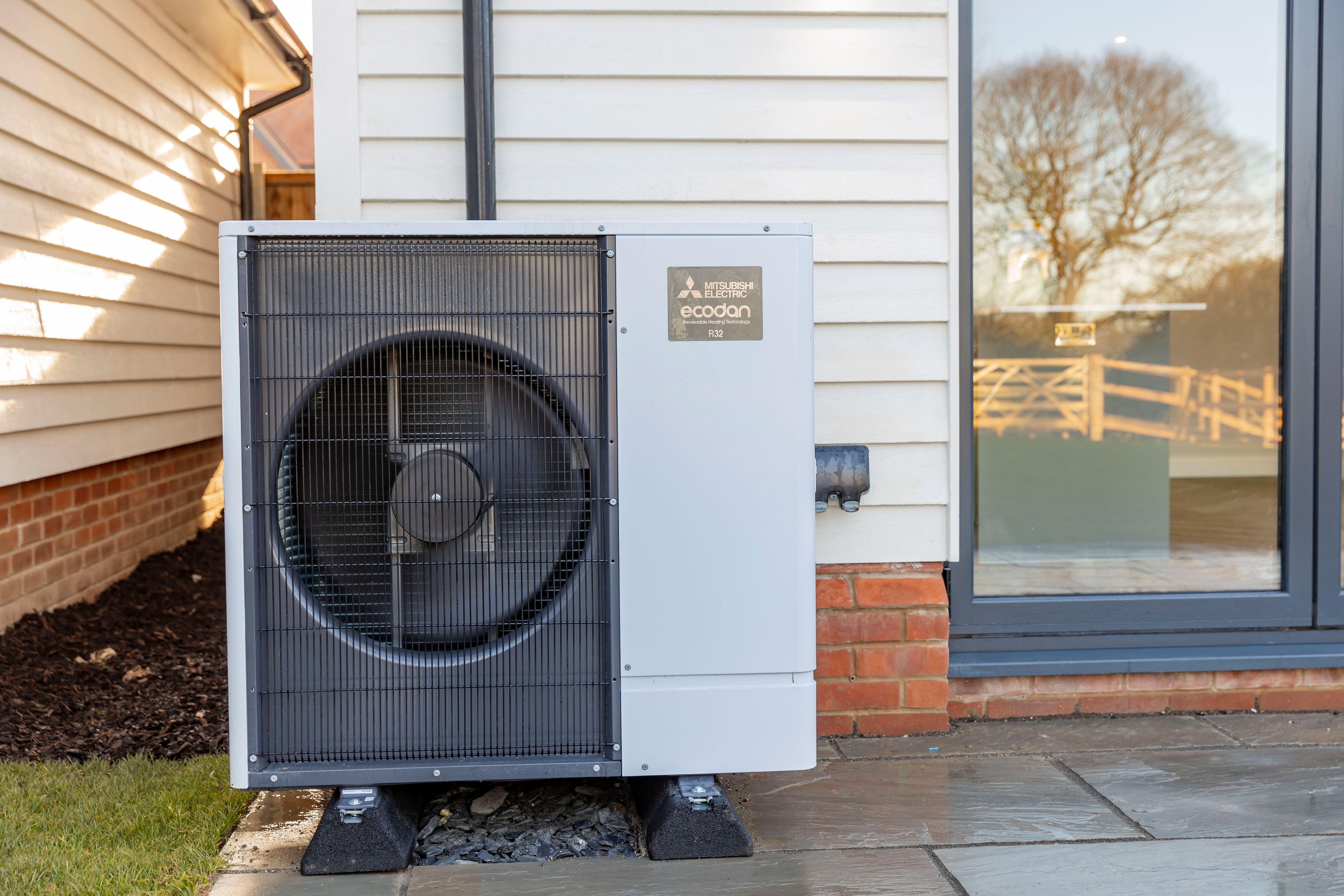 An air source heat pump standing on paving against the clapboard exterior of a house, near to a glass door