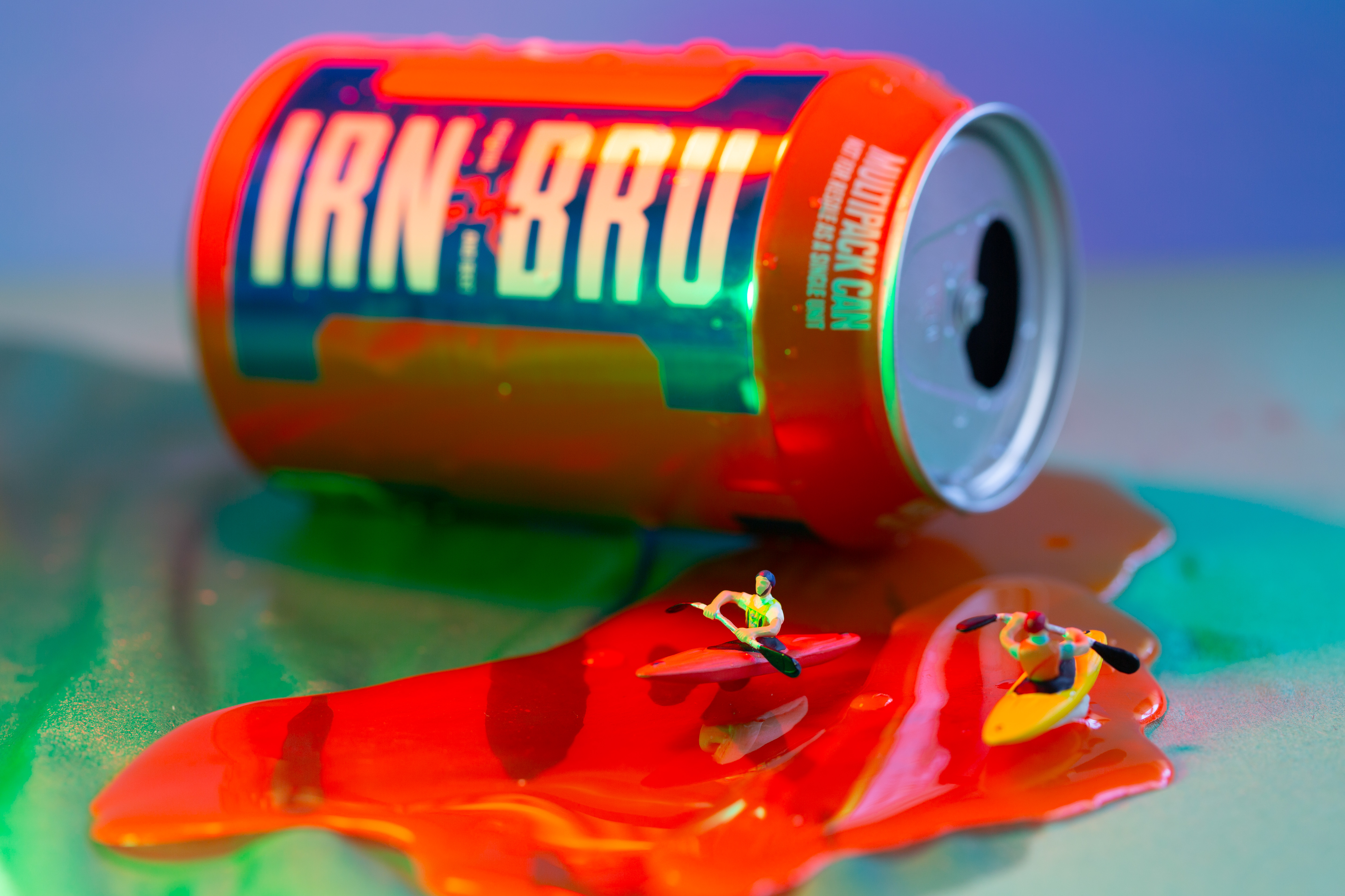Tiny figures in canoes on a puddle of orange liquid, next to a can of Irn-Bru