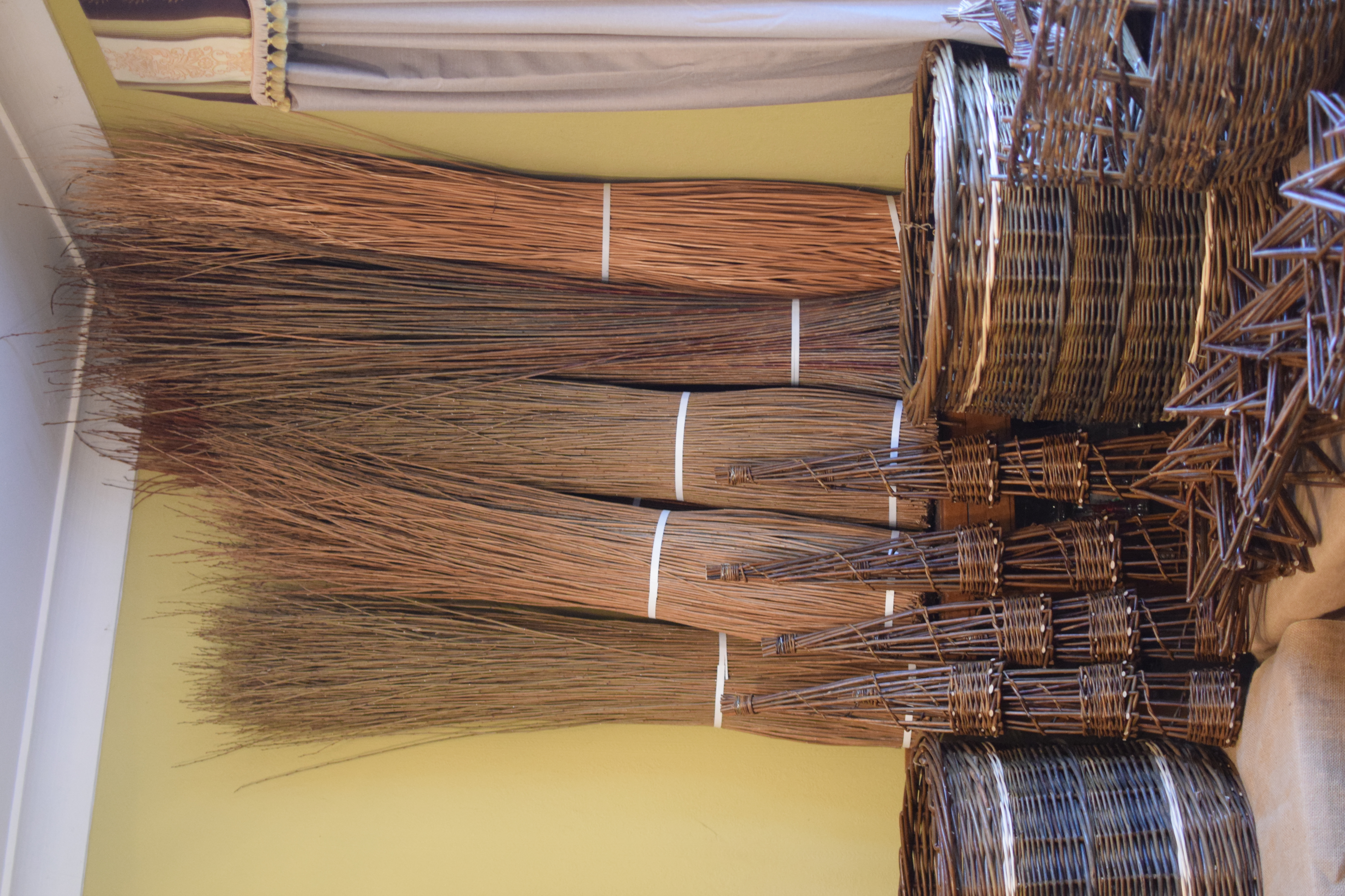 Large bales of willow leaning against a wall, with weaved baskets and other items sitting on the floor beside them