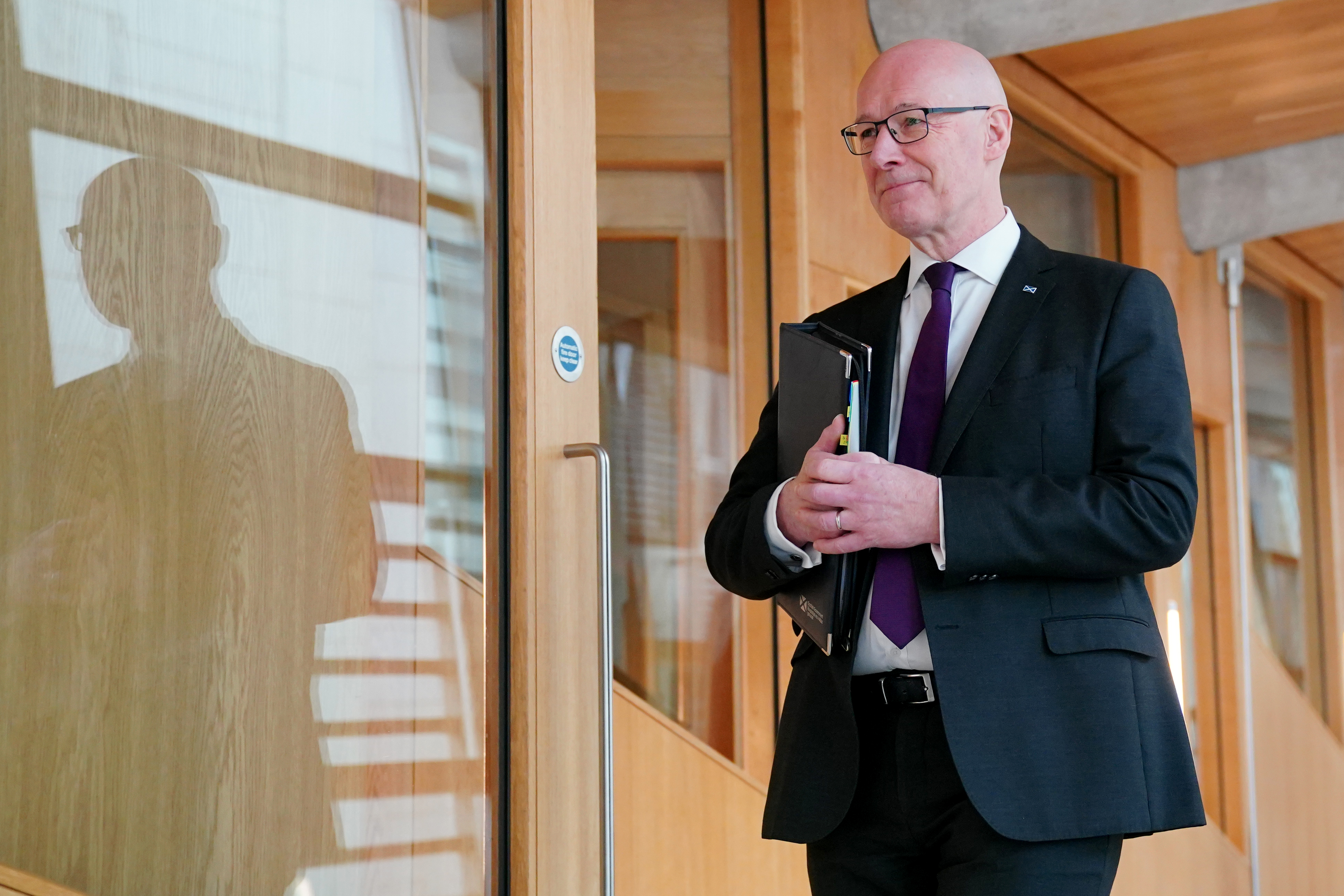 First Minister John Swinney smiles as he walks down a corridor in the Scottish Parliament