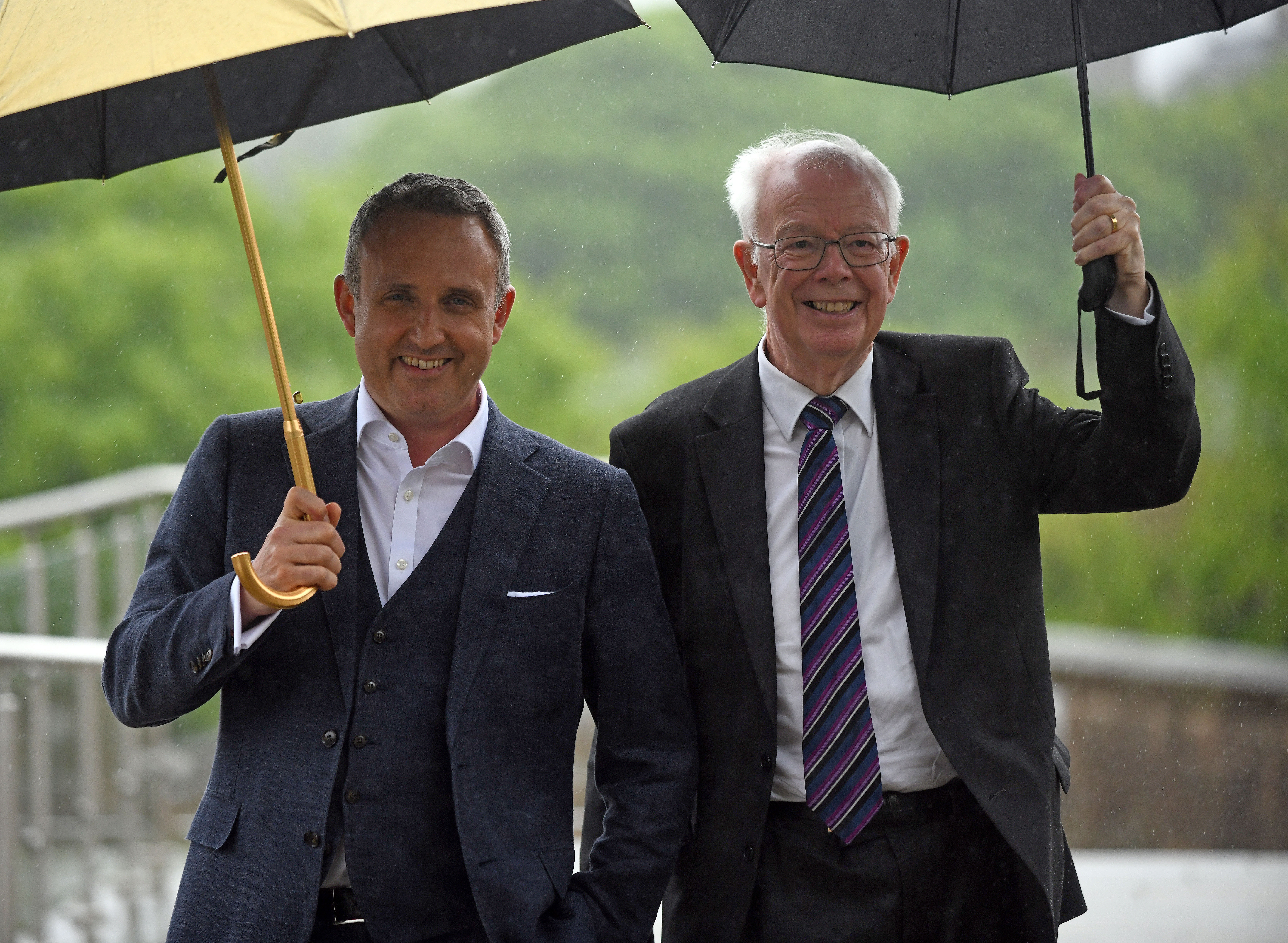 Alex Cole-Hamilton standing alongside Lord Wallace, with both men smiling and using umbrellas to shelter from the rain