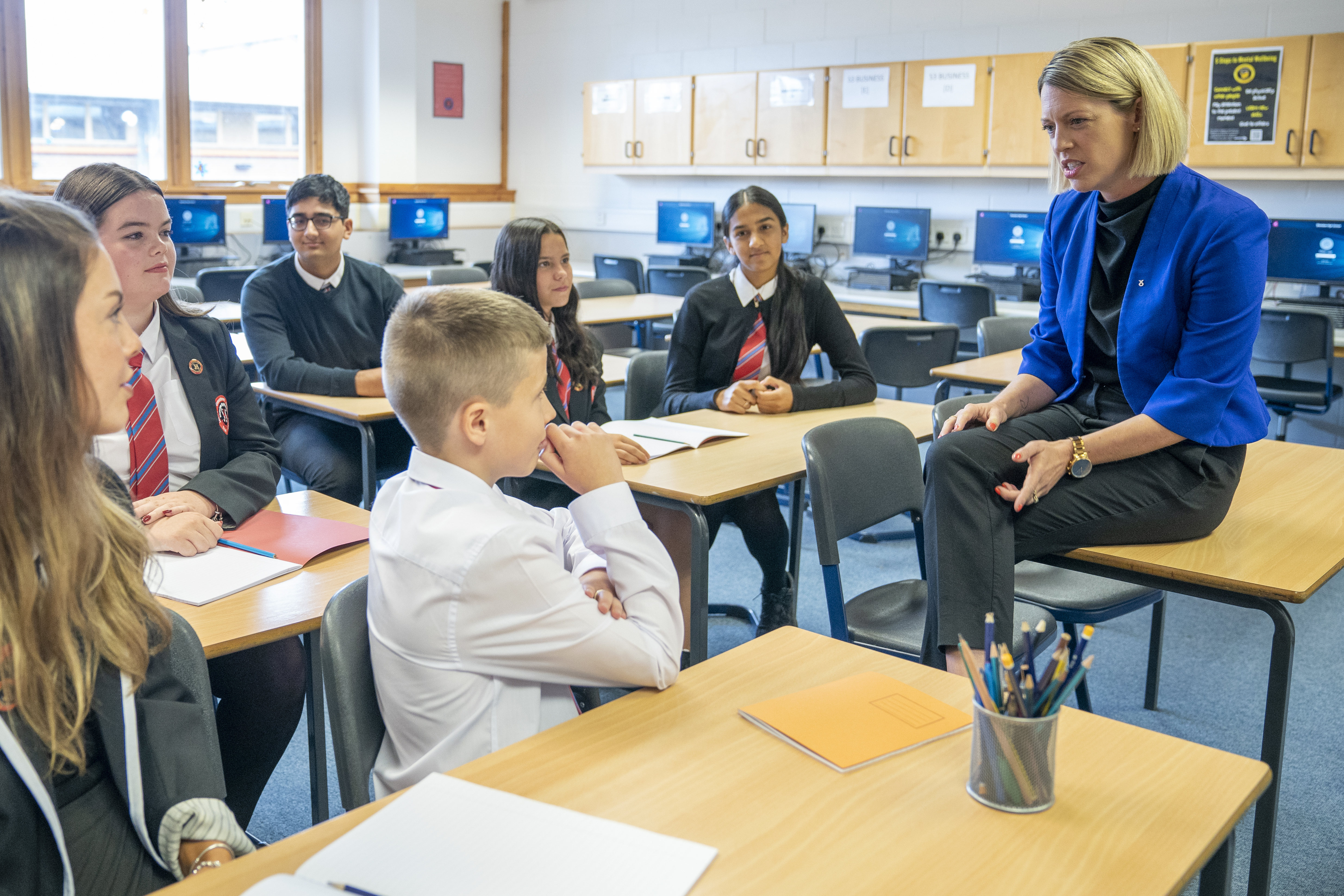 Jenny Gilruth sitting on a desk in a classroom while speaking to a group of senior school pupils