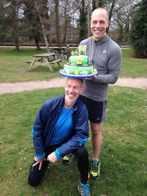 Darren Wood holding a cake celebrating his 500th parkrun with event founder Paul Sinton-Hewitt who is kneeling below the cake