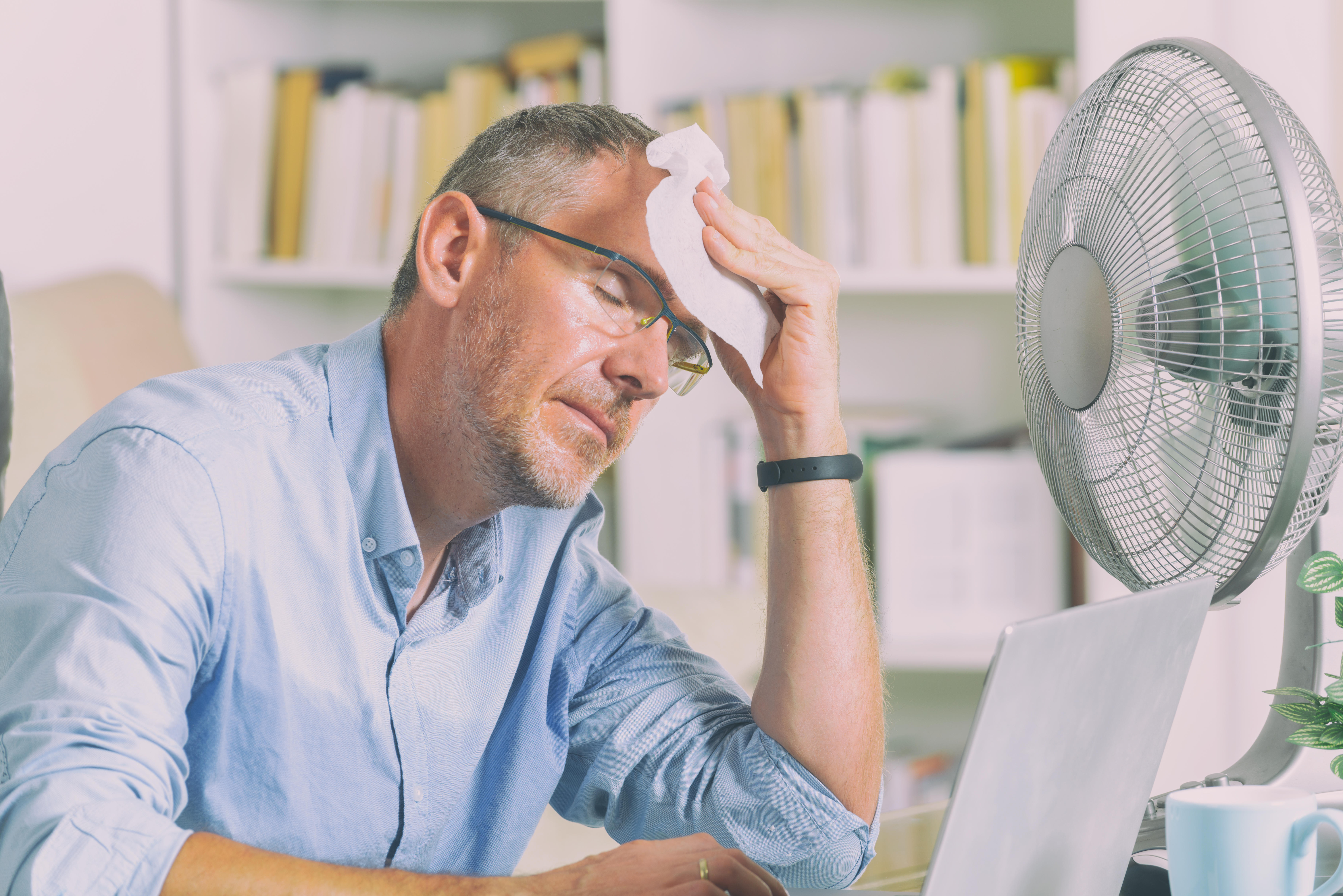 Man sitting in front of a fan in an office while wiping his forehead with sweat
