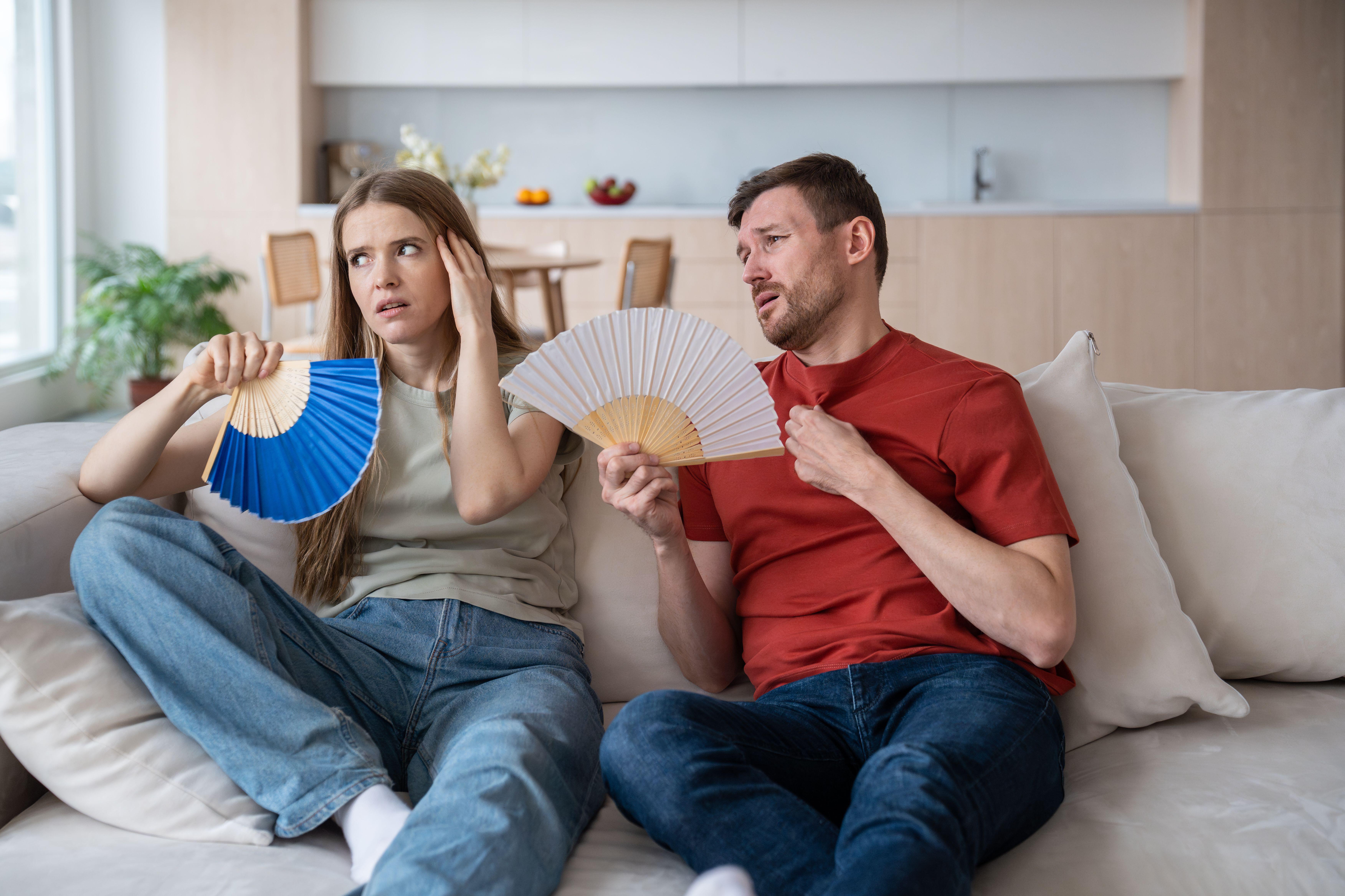 Overheated irritated married couple sitting on a sofa fanning themselves with fans