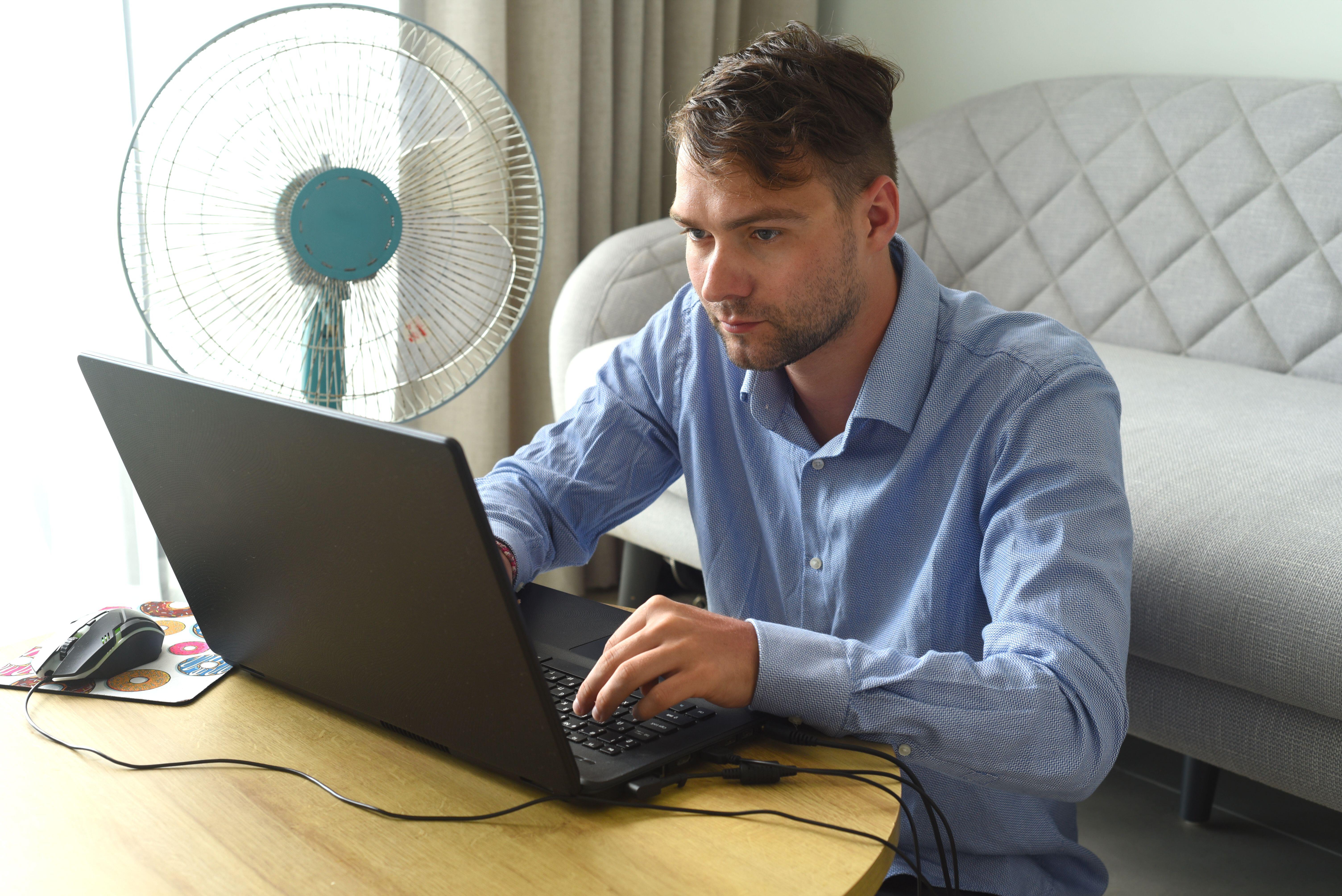 Young man working on a laptop at home next to a fan