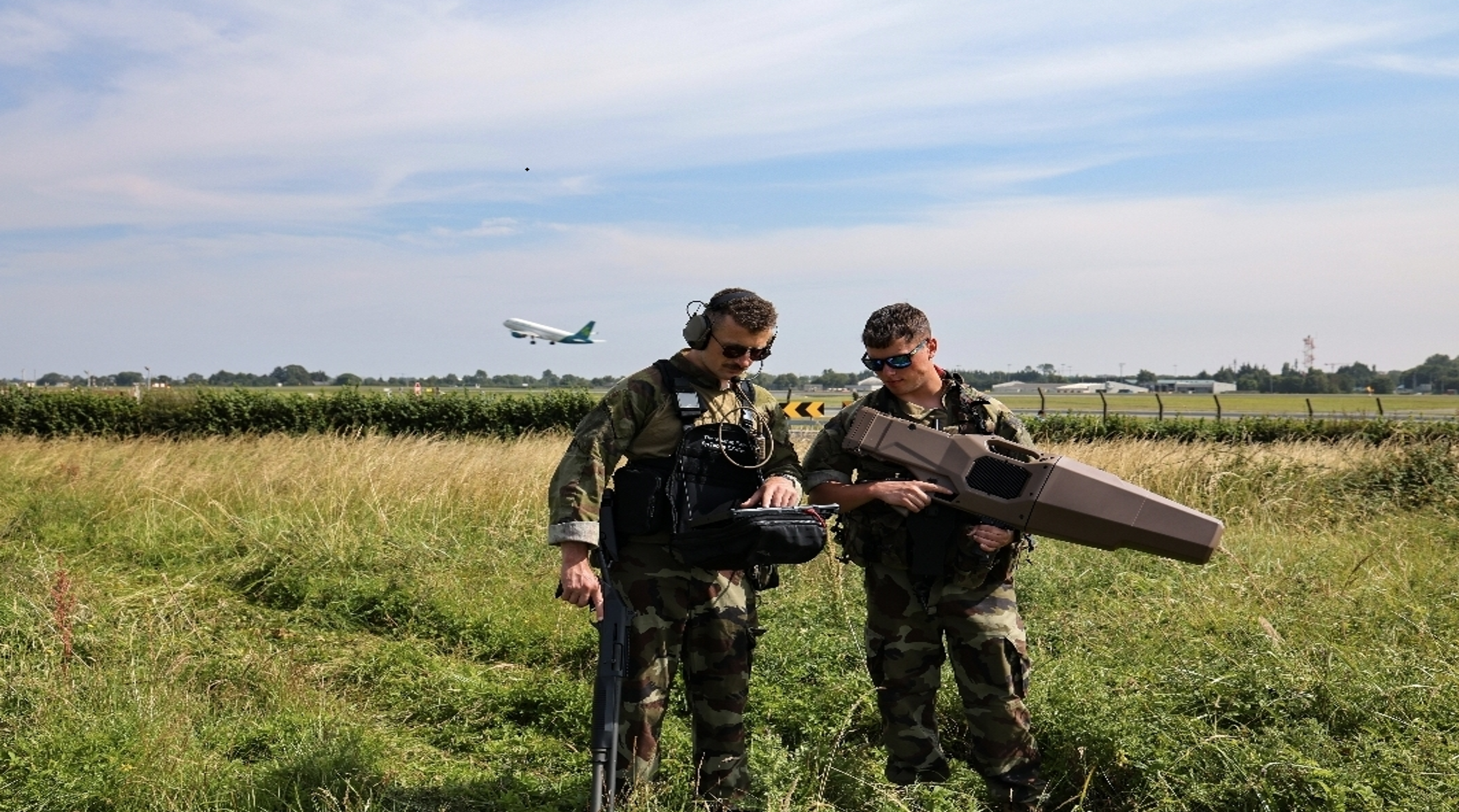 Two soldiers on grass using scanning equipment, as a plane takes off behind them