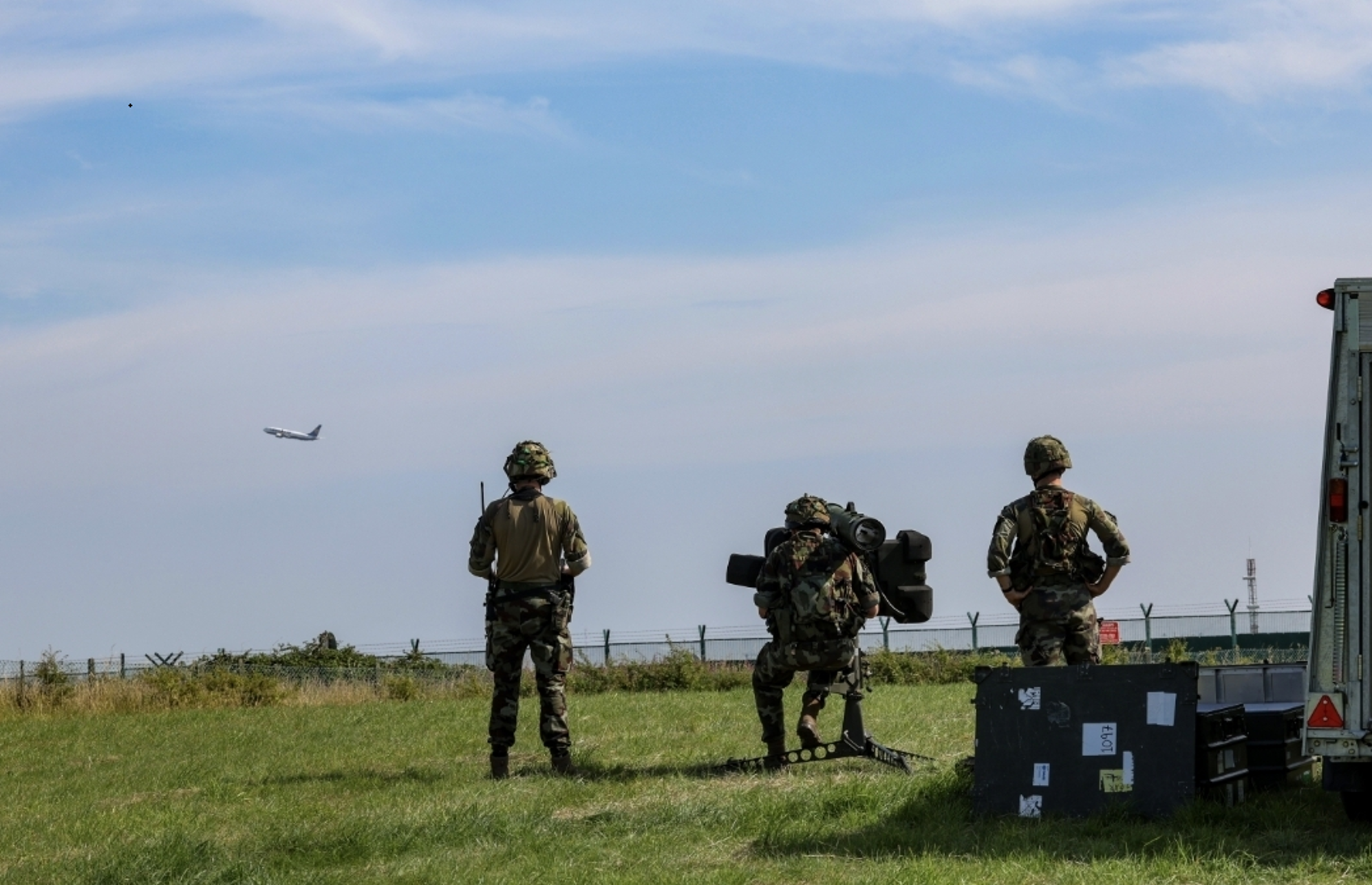 The backs of three soldiers on a grassy area, with one using a large piece of equipment, and a plane taking off in the distance