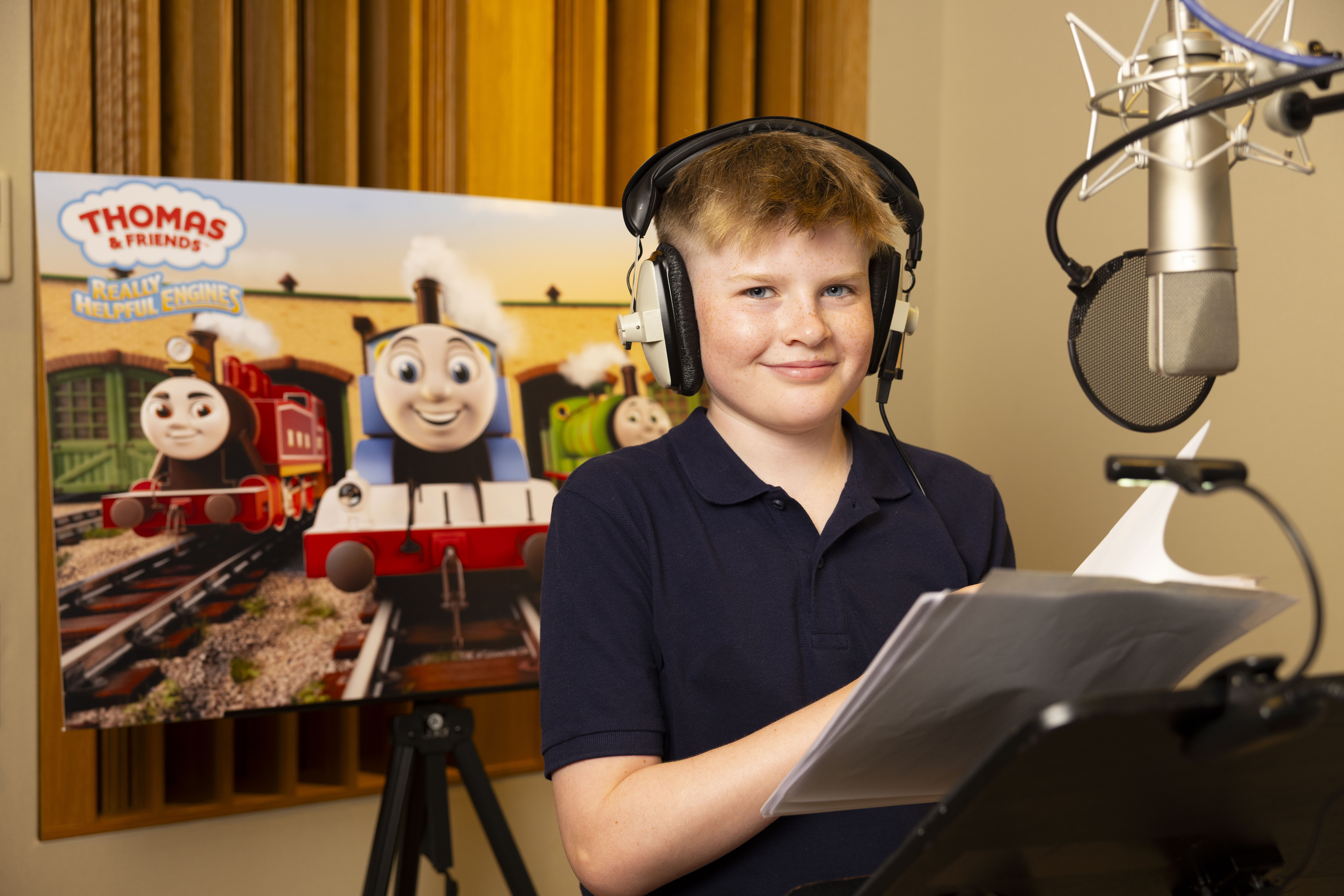 Harry Sparkes wearing a black polo and headphones standing in front of a microphone and Thomas The Tank Engine poster
