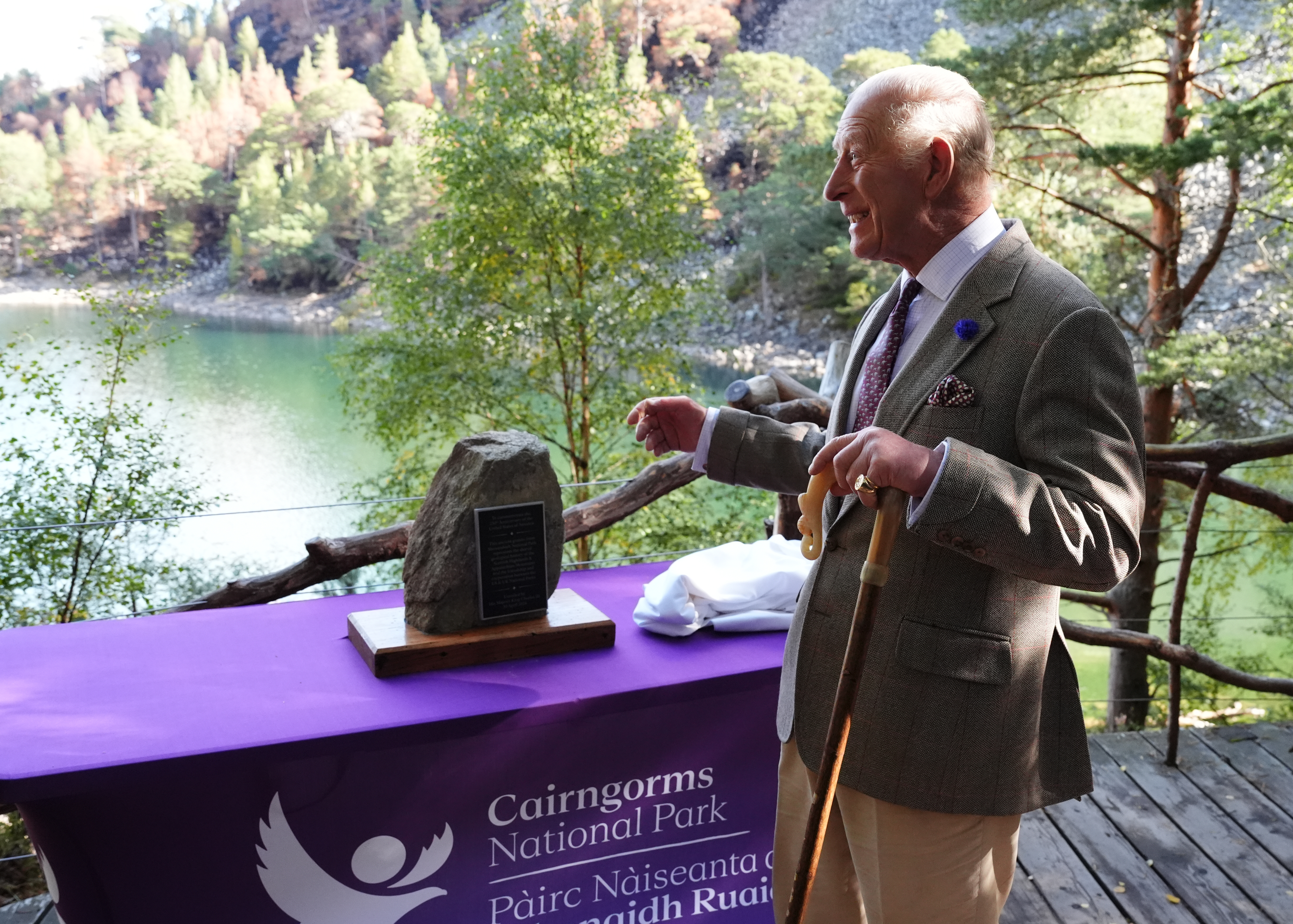 The King smiling, having unveiled a memorial stone featuring a plaque, which lies on a table above a loch