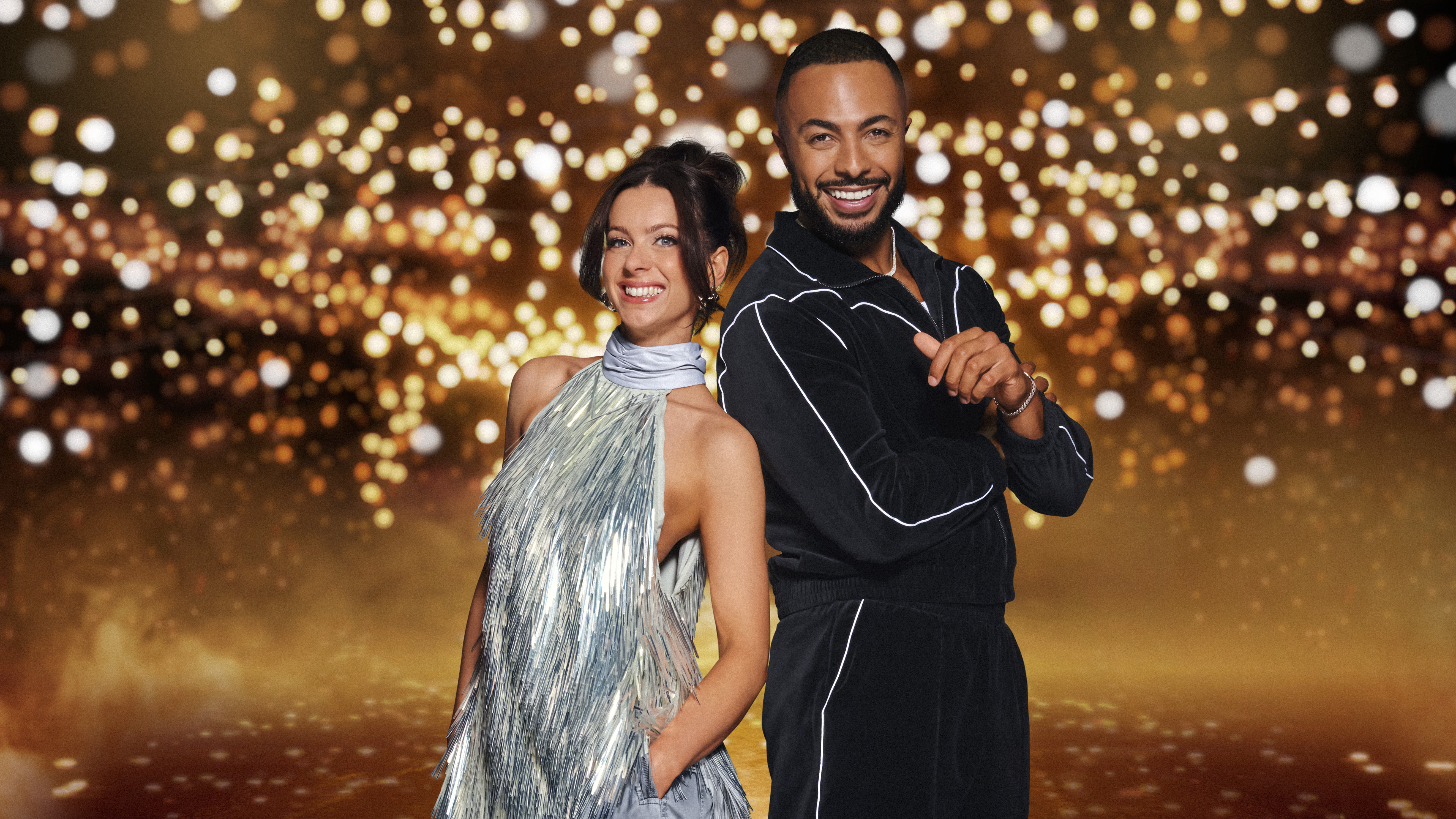 Abi Clarke wearing a silver halter neck dress and Tyler West raises his arms to his chest smiling wearing in a black jumpsuit