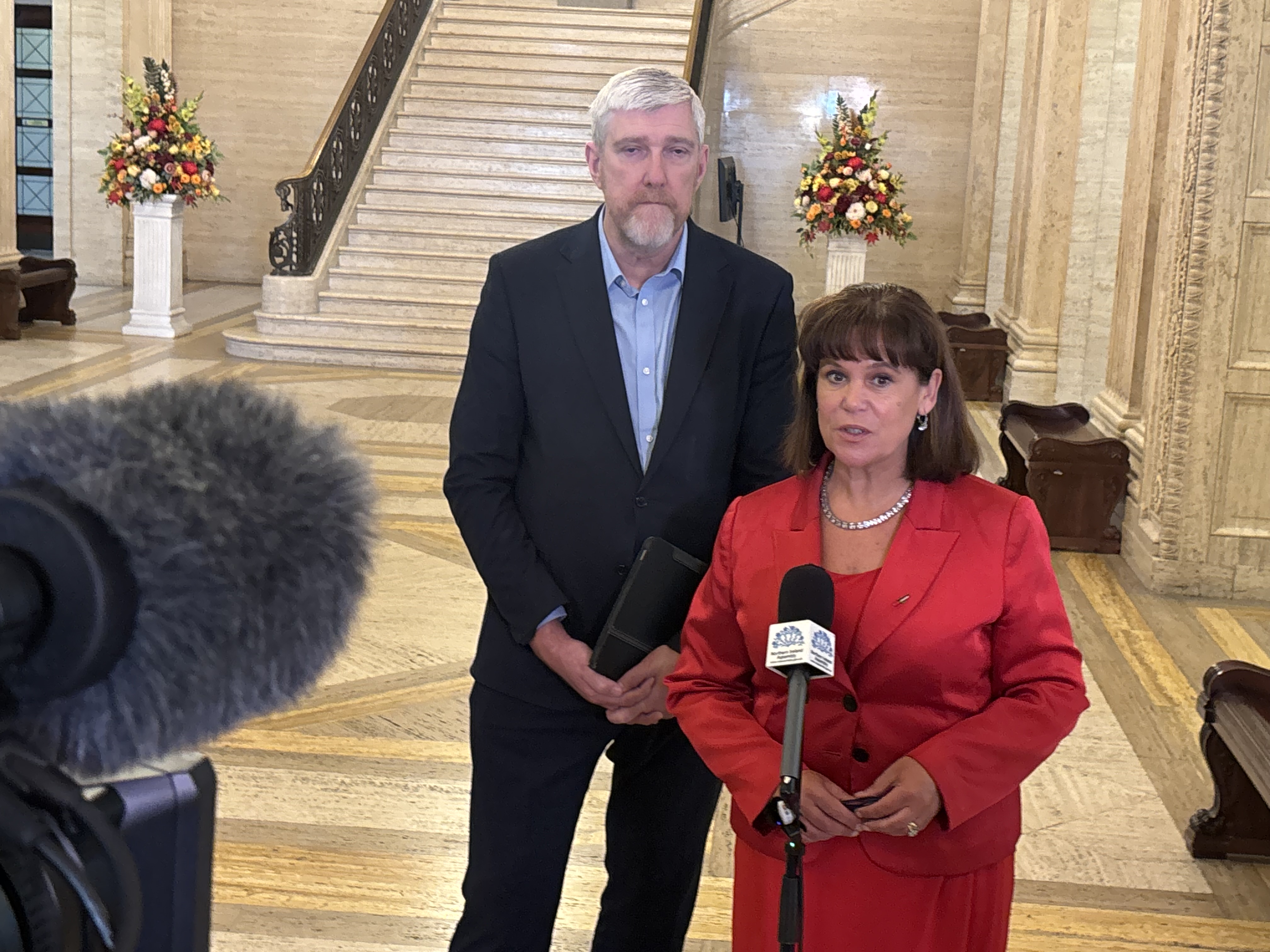 Mary-Lou McDonald speaking into a microphone in Parliament Buildings, with John O'Dowd behind her