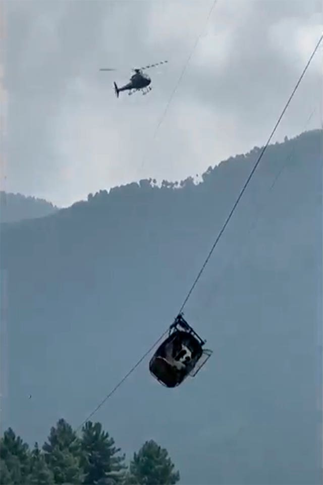 The cable car dangles hundreds of feet above the ground while it is circled by a helicopter