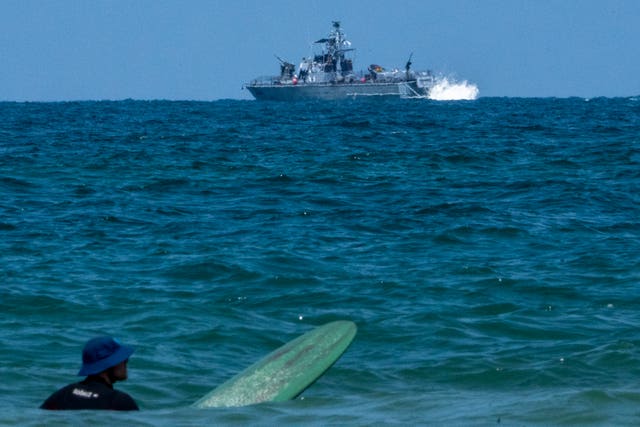 A surfer waits for waves while an Israeli military naval ship patrols the Mediterranean Sea off the coast of Hadera, Israel 