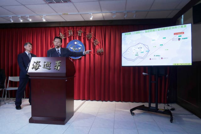 Two men stand at a lectern, there's a red curtain behind them all the way around the wall and to the right is a large TV screen showing a graphic of a map