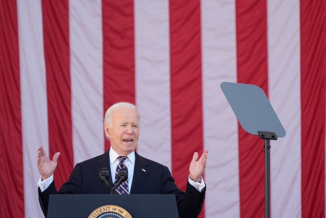 President Joe Biden raises his hands while speaking at Arlington National Cemetery, with an American flag in the background