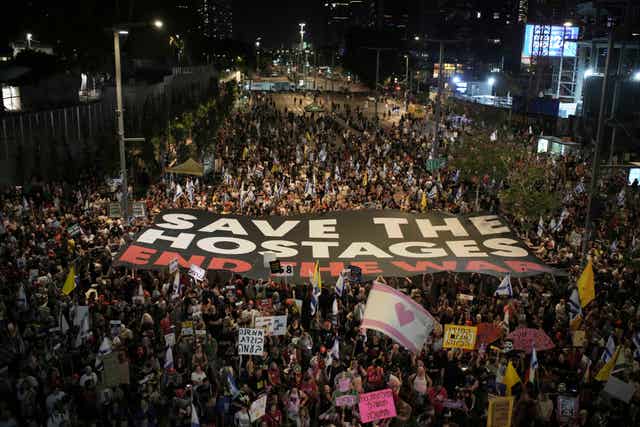A huge crowd of people carrying a large banner which reads 'Save the hostages, end the war'