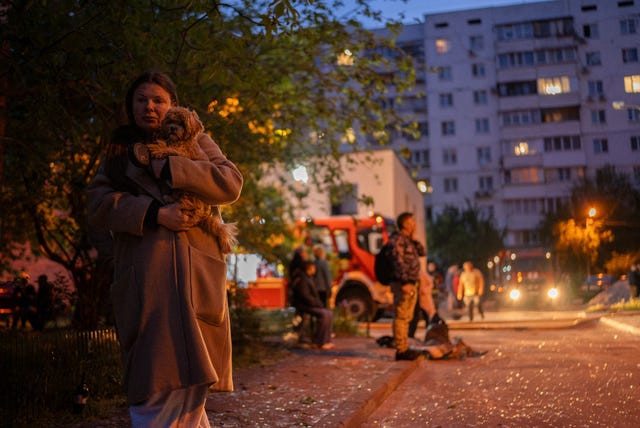 A woman carries her dog as she walks on a street with pieces of broken glass at the site of a residential building