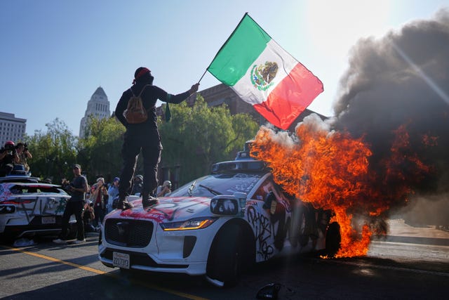 A protester stands on a burning Waymo taxi