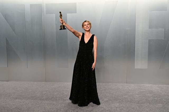 Jessie Buckley, wearing a black dress, holds her Academy Award in the air with her right hand