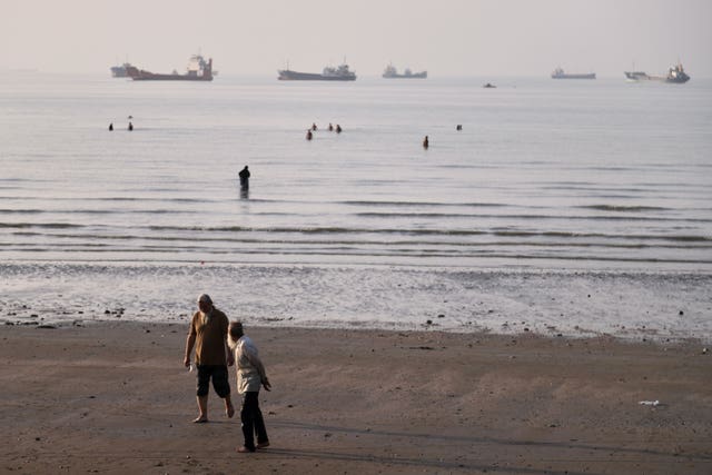 Men walk on the shoreline as people wade in the water in the Strait of Hormuz, where cargo and service vessels are anchored off Bandar Abbas, Iran