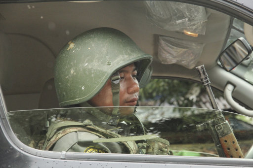 A heavily militarised police officer sits in a car, wearing a hat and holding a rifle