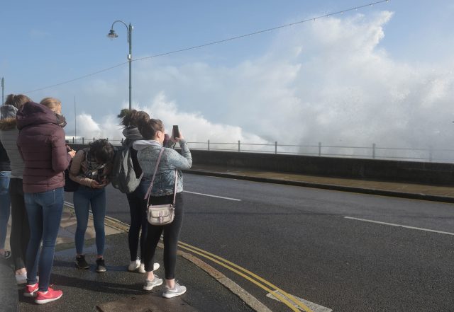 Members of the public take photographs of waves crashing on the sea wall at Penzanze