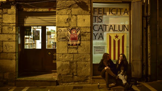 Two women seated on a step in front of an ''estelada'' or Catalonia independence flag reading, ''Congratulations Catalonia''